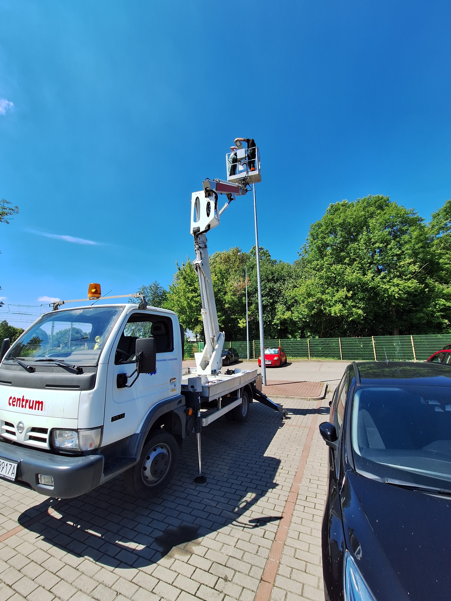 Biały samochód z podnośnikiem koszowym na parkingu, dwóch pracowników w koszu na wysokości, błękitne niebo w tle.