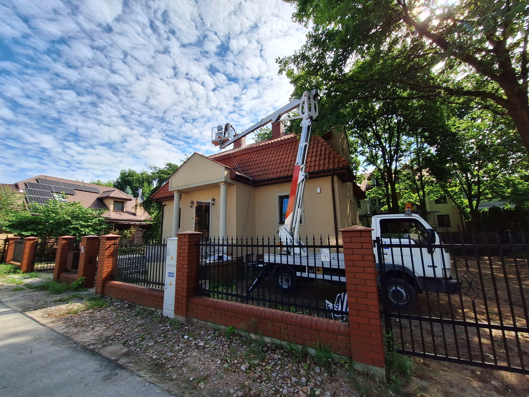 Podnośnik koszowy ustawiony przy domu jednorodzinnym w Karczewie, widoczny zza ogrodzenia. Kosz wysięgnika sięga dachu budynku.