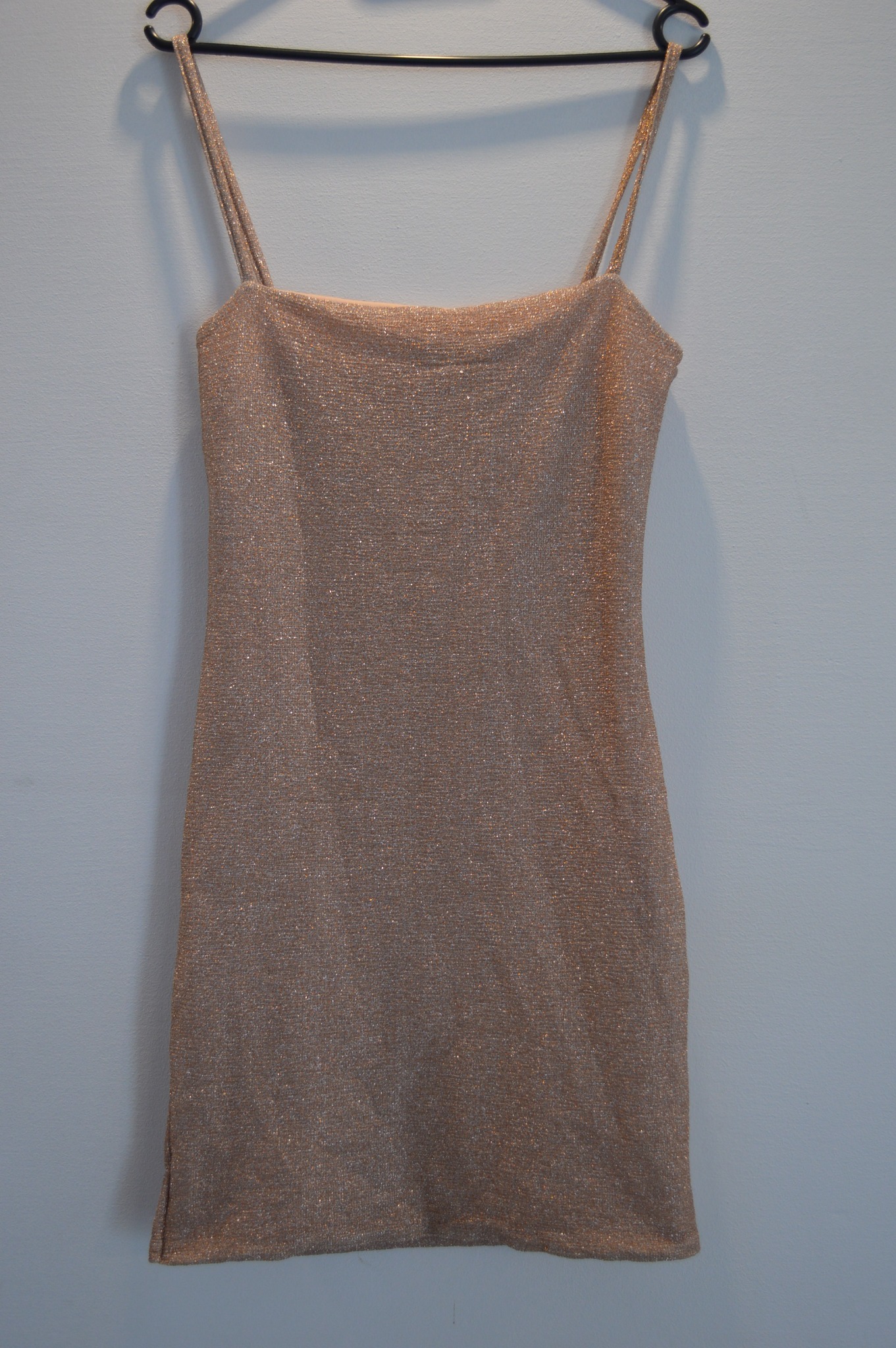 Krótka, połyskująca sukienka na ramiączkach, wisząca na czarnym wieszaku na tle szarej ściany.