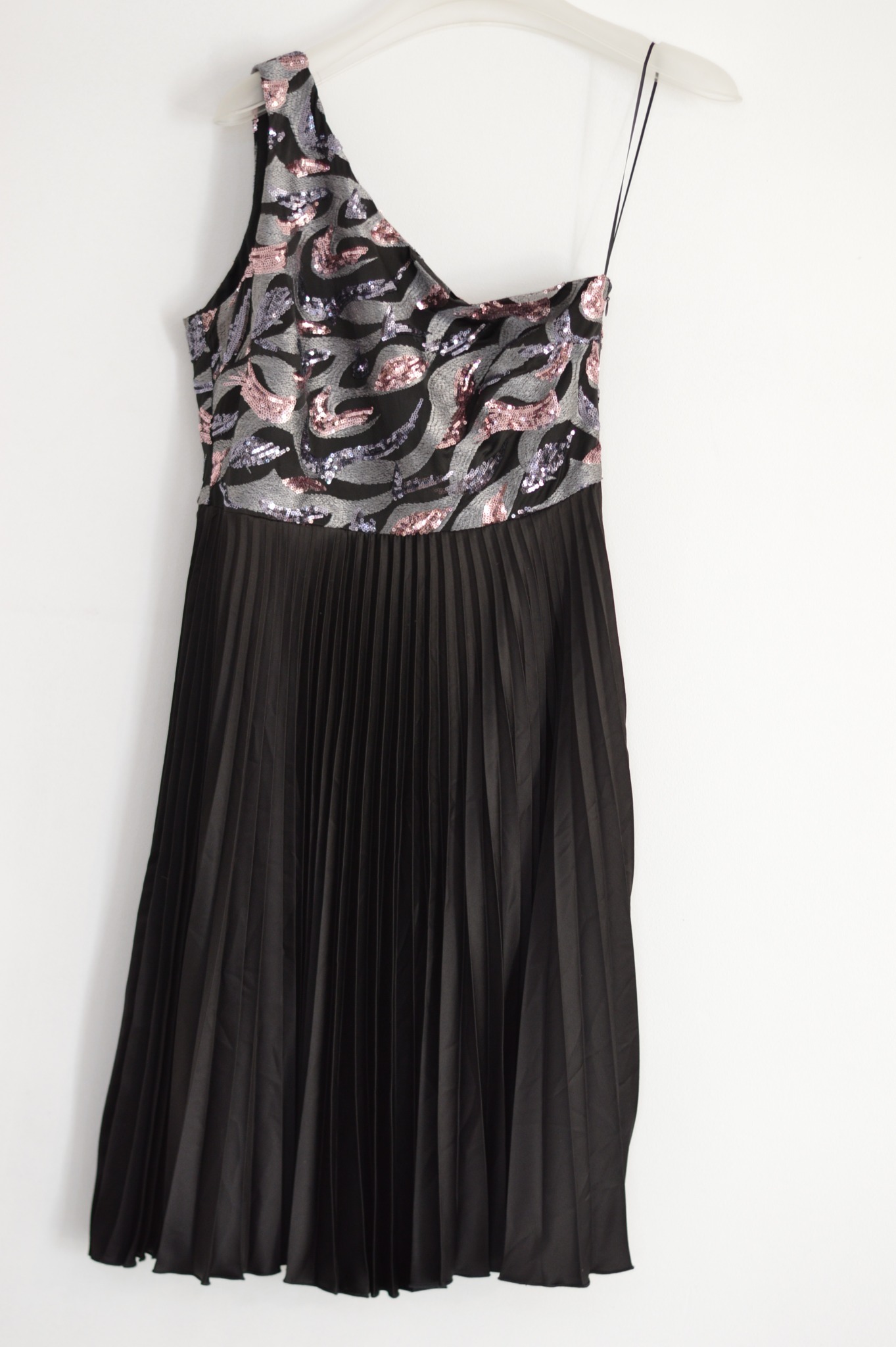 Czarna plisowana sukienka na jedno ramię z cekinową górą w odcieniach różu i srebra, wisząca na białym wieszaku na tle białej ściany.