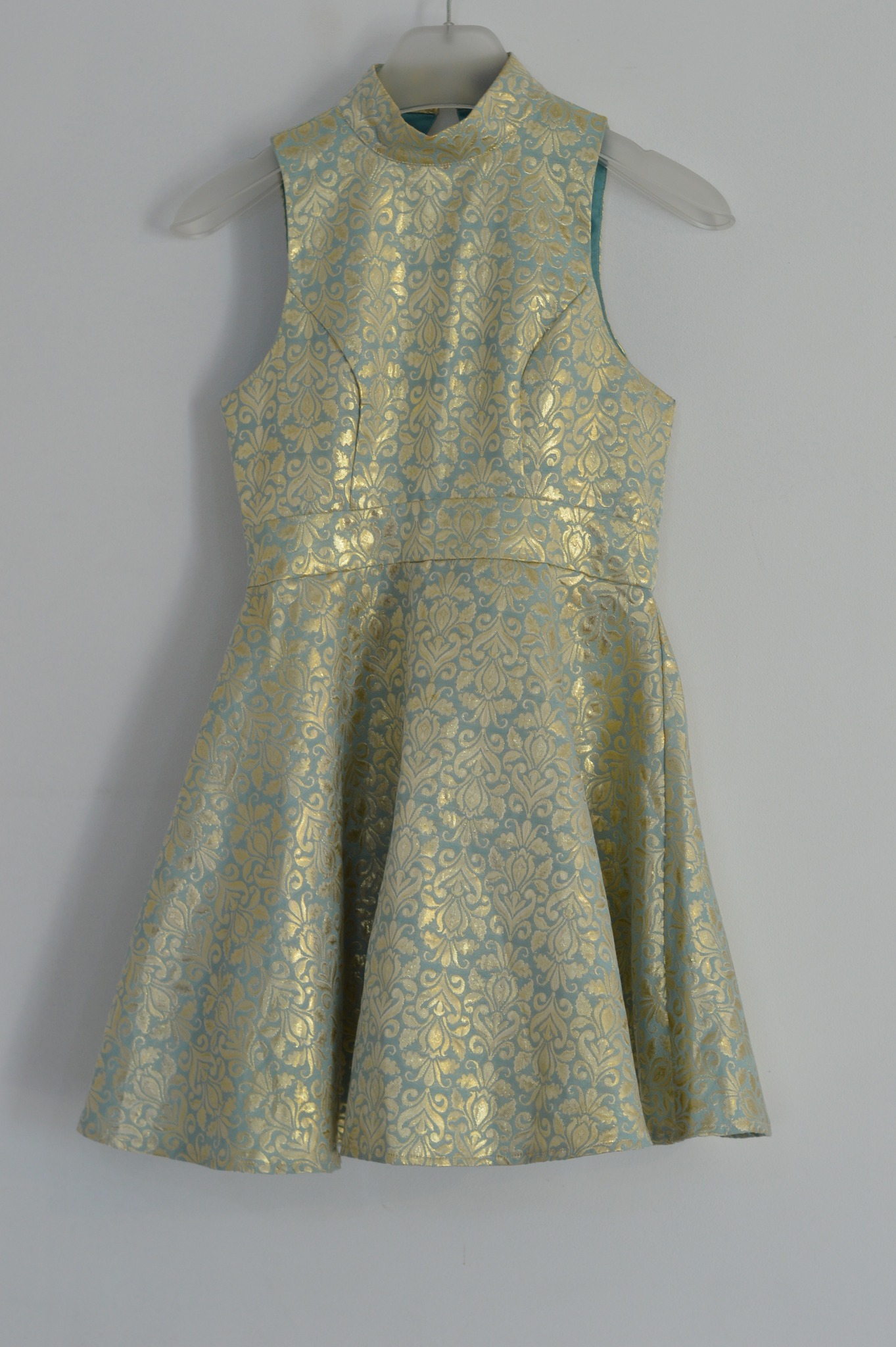 Sukienka bez rękawów z wysokim kołnierzem, w kolorze miętowym z wytłaczanym złotym wzorem, wisząca na plastikowym wieszaku na tle białej ściany.