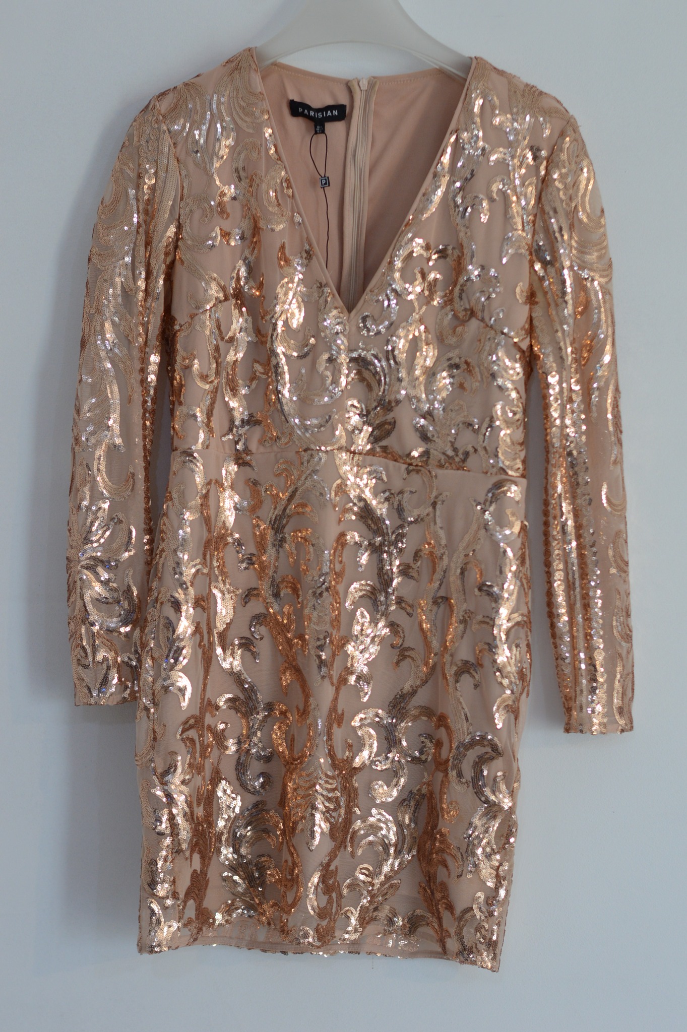Sukienka koktajlowa mini, beżowa, z długimi rękawami, pokryta wzorem z błyszczących, złotych cekinów, wisząca na wieszaku na tle białej ściany.