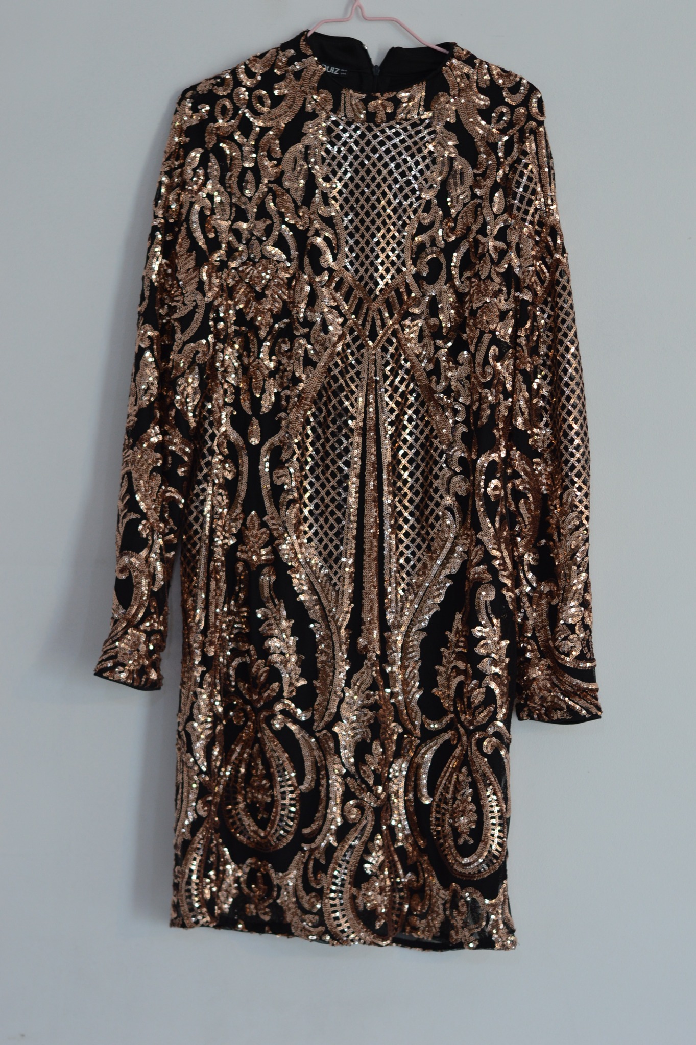 Czarna sukienka koktajlowa z długim rękawem, pokryta bogatym, złotym, cekinowym wzorem w stylu art deco, wisząca na różowym wieszaku na tle białej ściany.