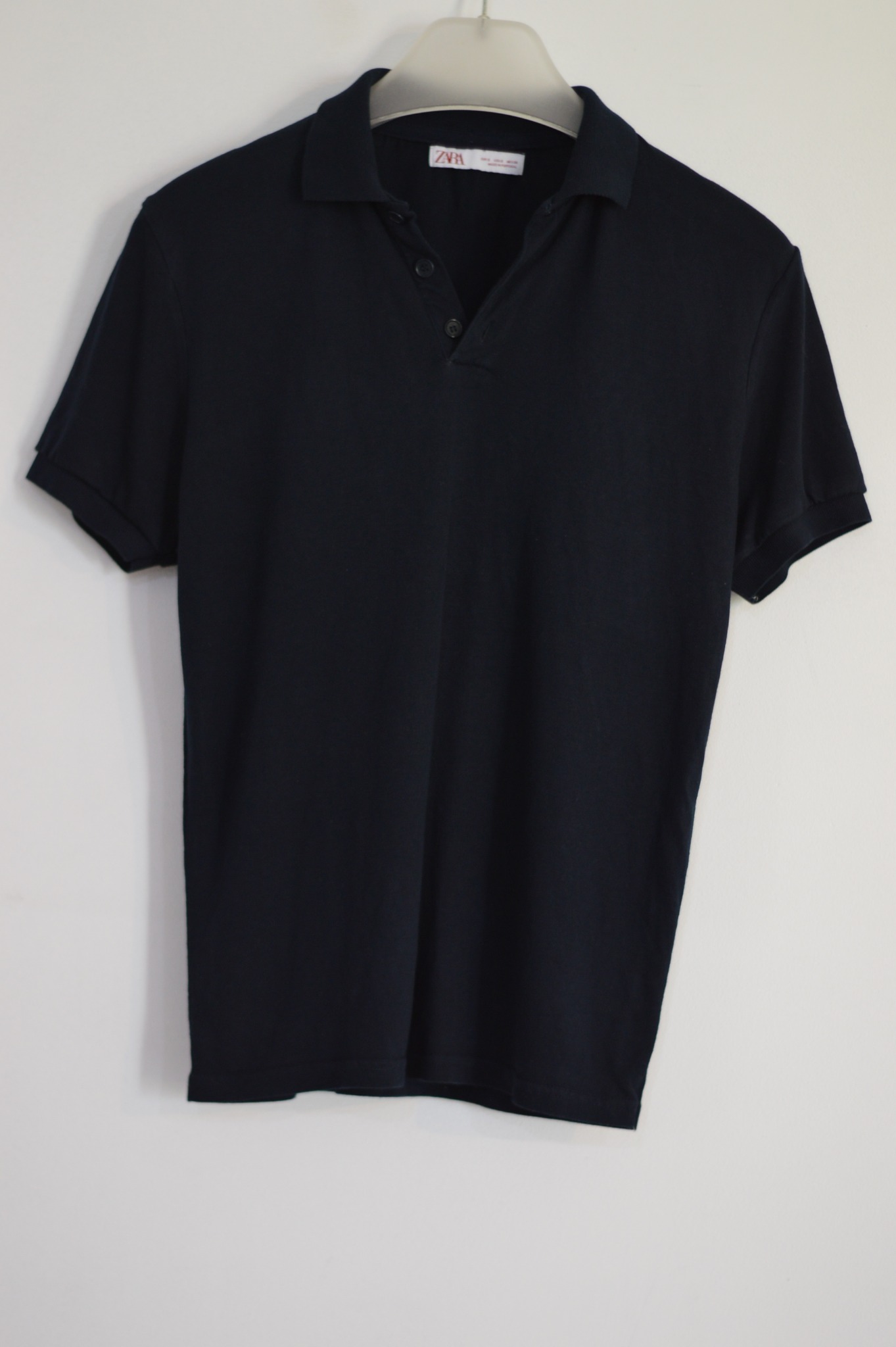Granatowa koszulka polo marki ZARA, zawieszona na plastikowym wieszaku na tle białej ściany, widoczne trzy guziki i krótki rękaw.