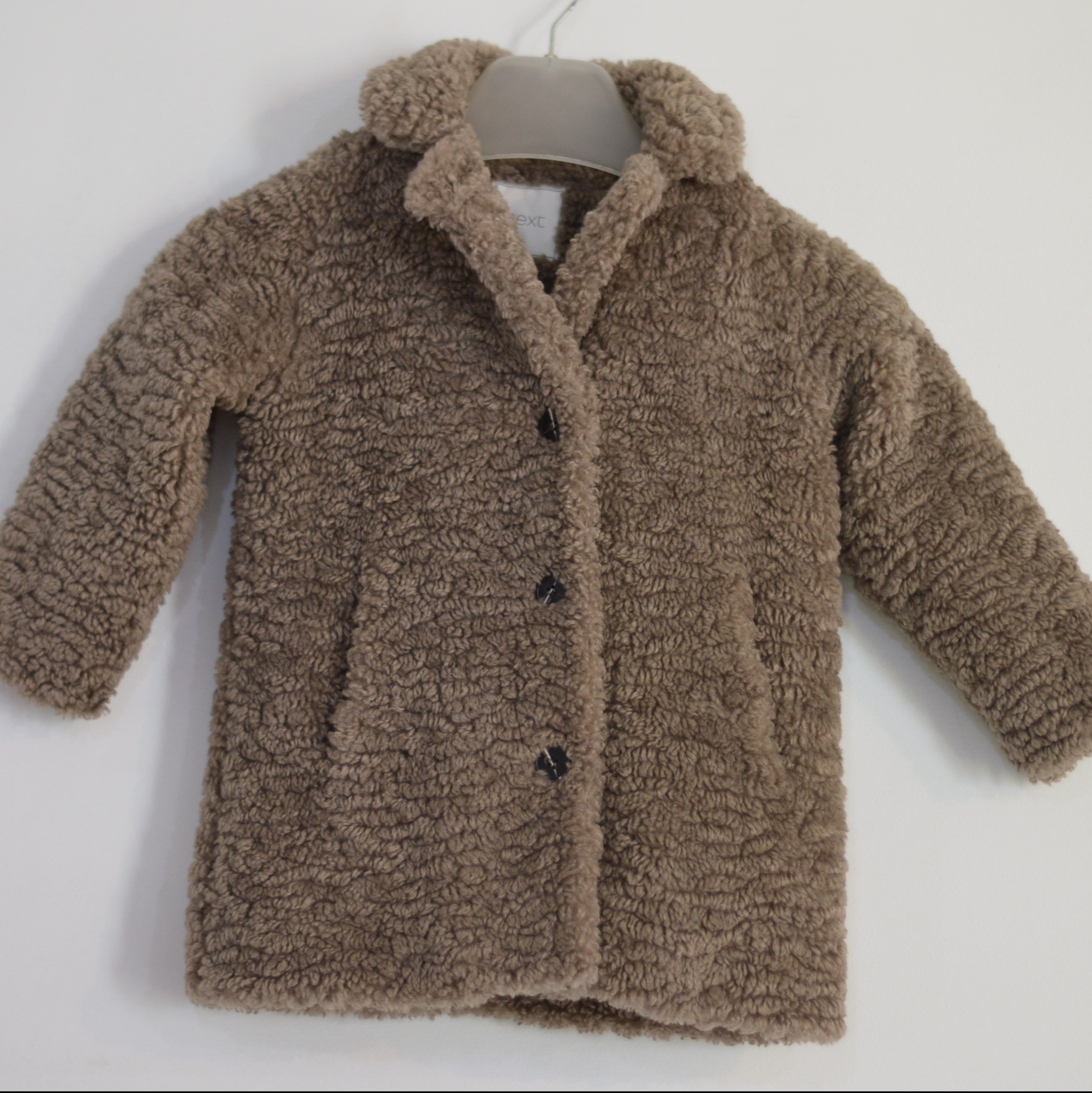 Jasnobrązowa kurtka z imitacji baranka, zapinana na trzy czarne guziki, wisząca na szarym wieszaku na białym tle. Widoczna metka marki 'Next'.