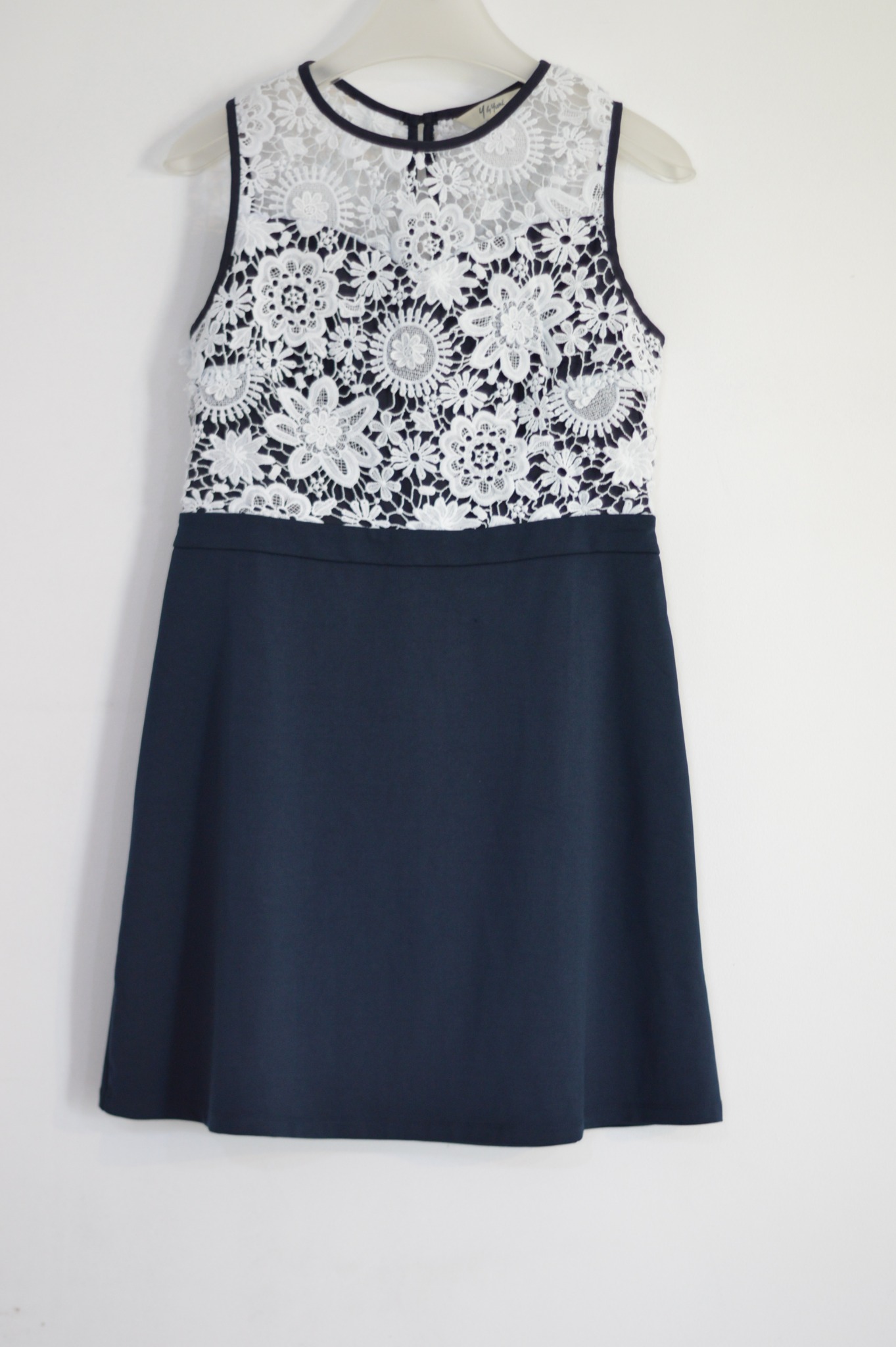 Granatowa sukienka z białą koronkową górą, wisząca na wieszaku na tle białej ściany.