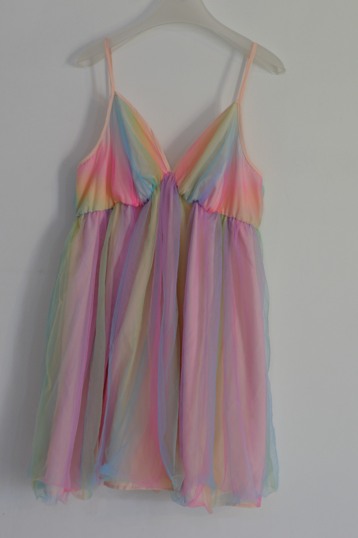 Sukienka na ramiączkach w pastelowych kolorach tęczy, wisząca na plastikowym wieszaku na białej ścianie.