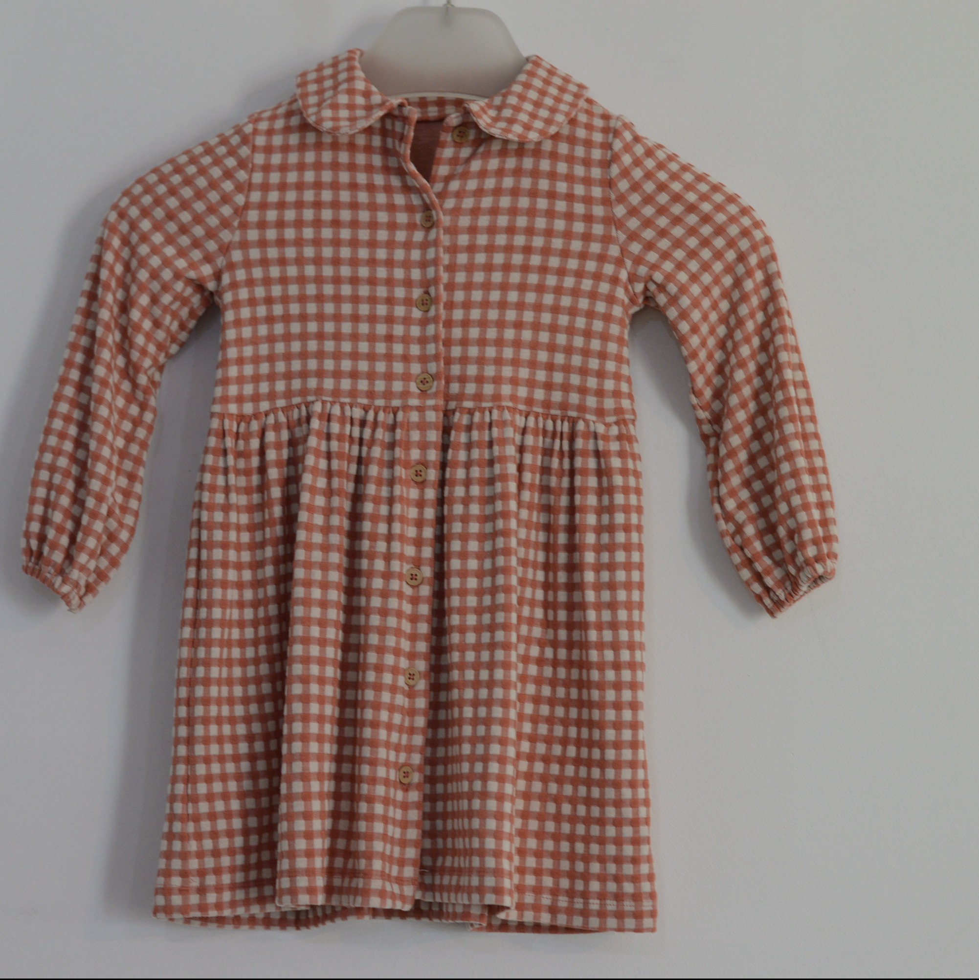 Dziewczęca sukienka w drobną kratkę, w odcieniach pomarańczowo-kremowych, z kołnierzykiem i zapinana na guziki, wisząca na wieszaku na tle jasnej ściany.