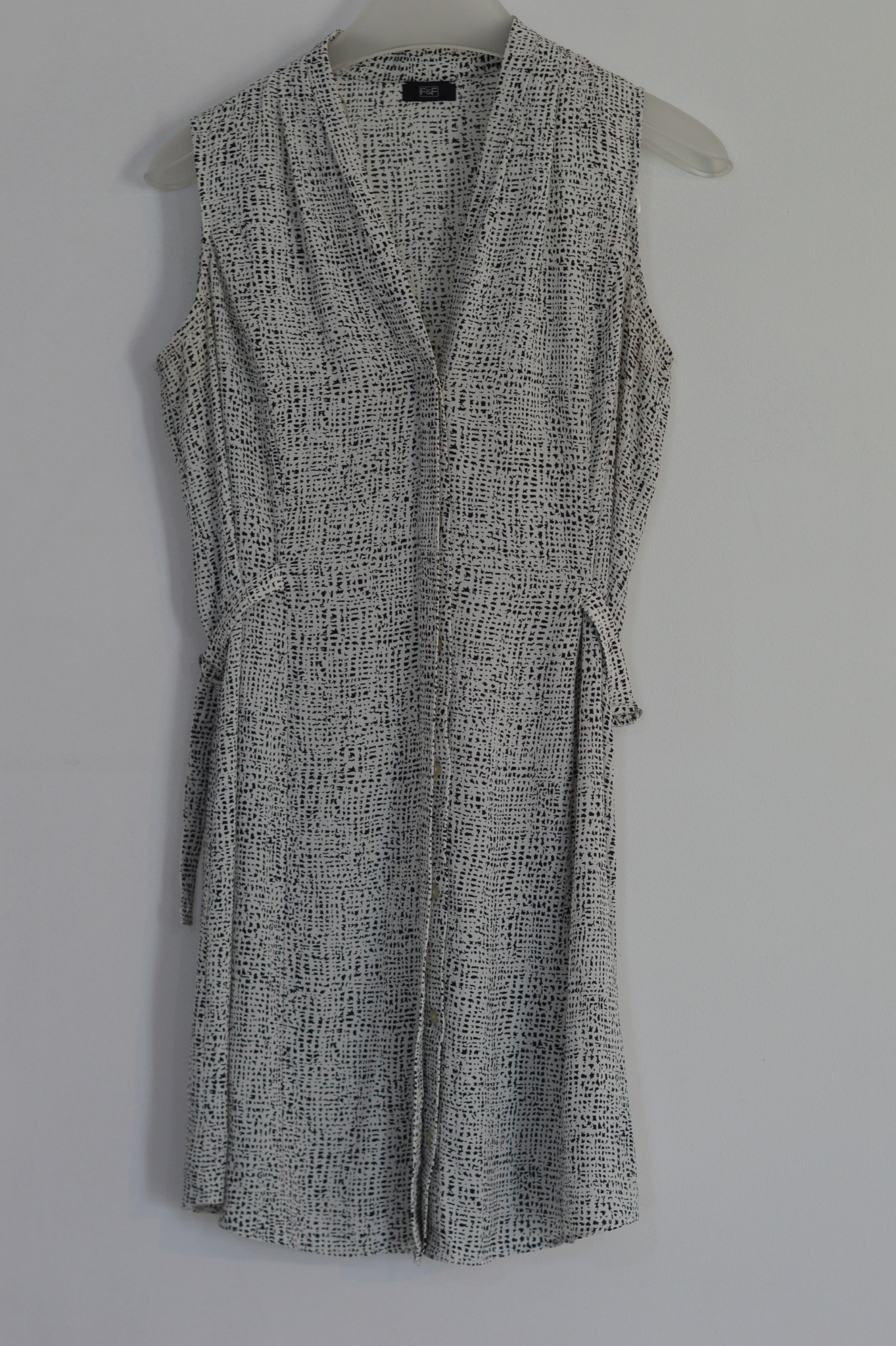 Damska, letnia sukienka bez rękawów w drobny wzór, wisząca na plastikowym wieszaku na tle białej ściany.