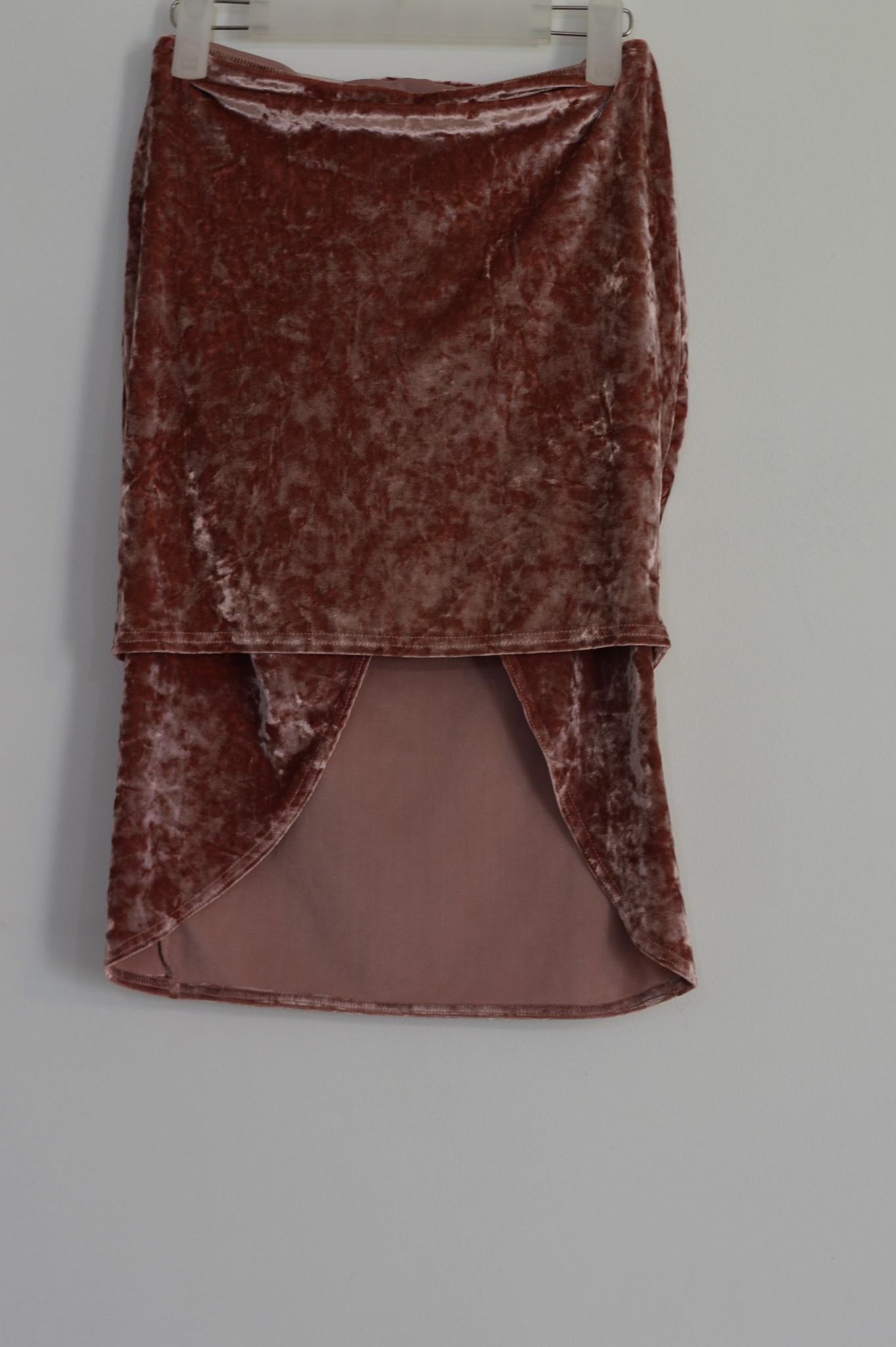 Różowa, aksamitna spódnica z asymetrycznym dołem, wisząca na wieszaku na tle szarej ściany.