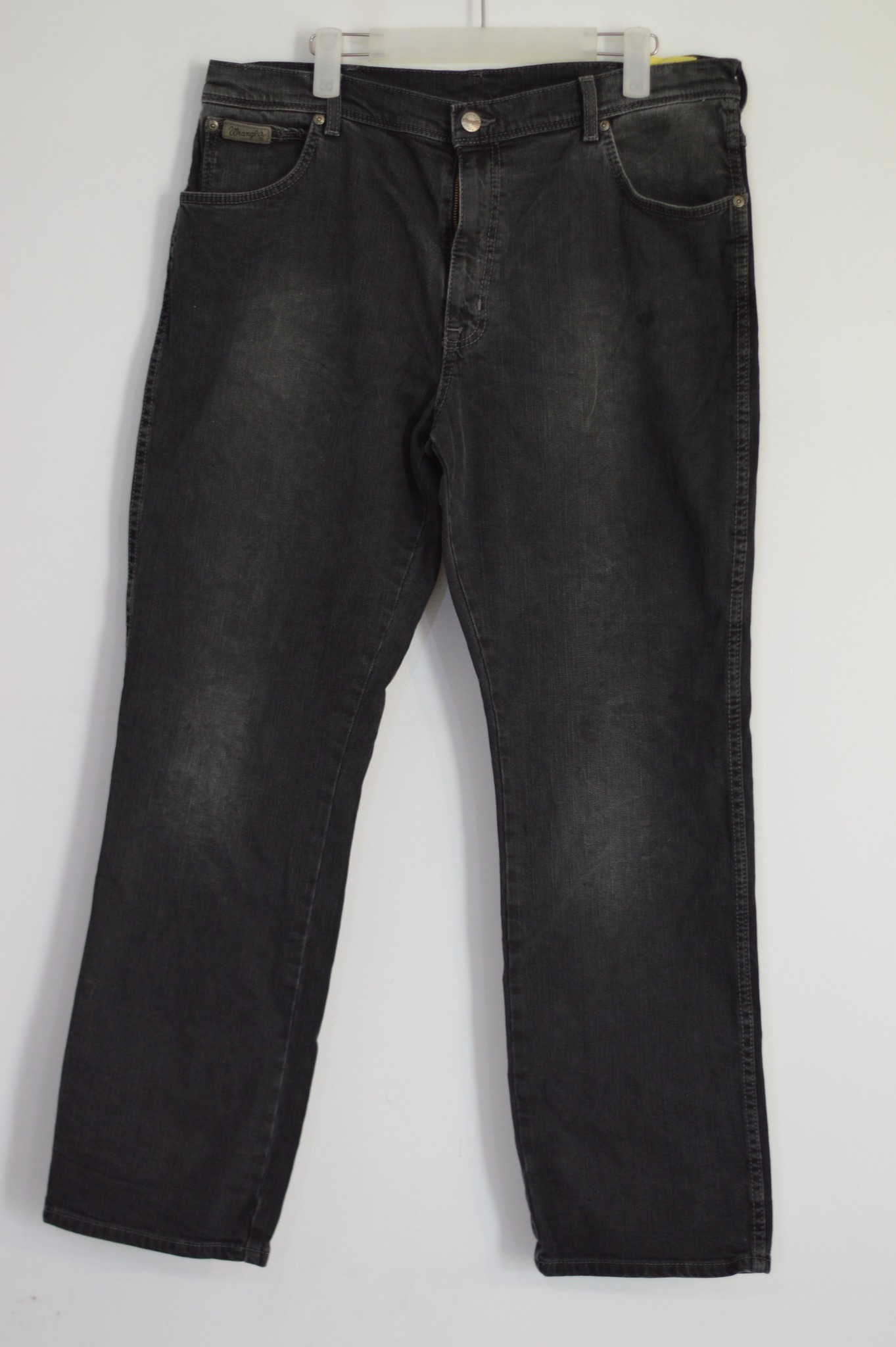 Czarne jeansy Wrangler, widoczne na wieszaku, z przetarciami i metalowymi elementami, na białym tle.