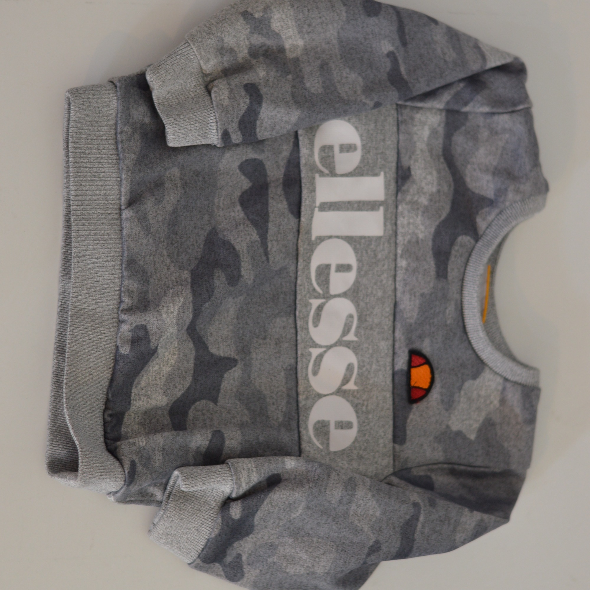 Szara bluza dresowa marki Ellesse z kamuflażem, widoczne logo na przodzie i ściągacze przy rękawach.