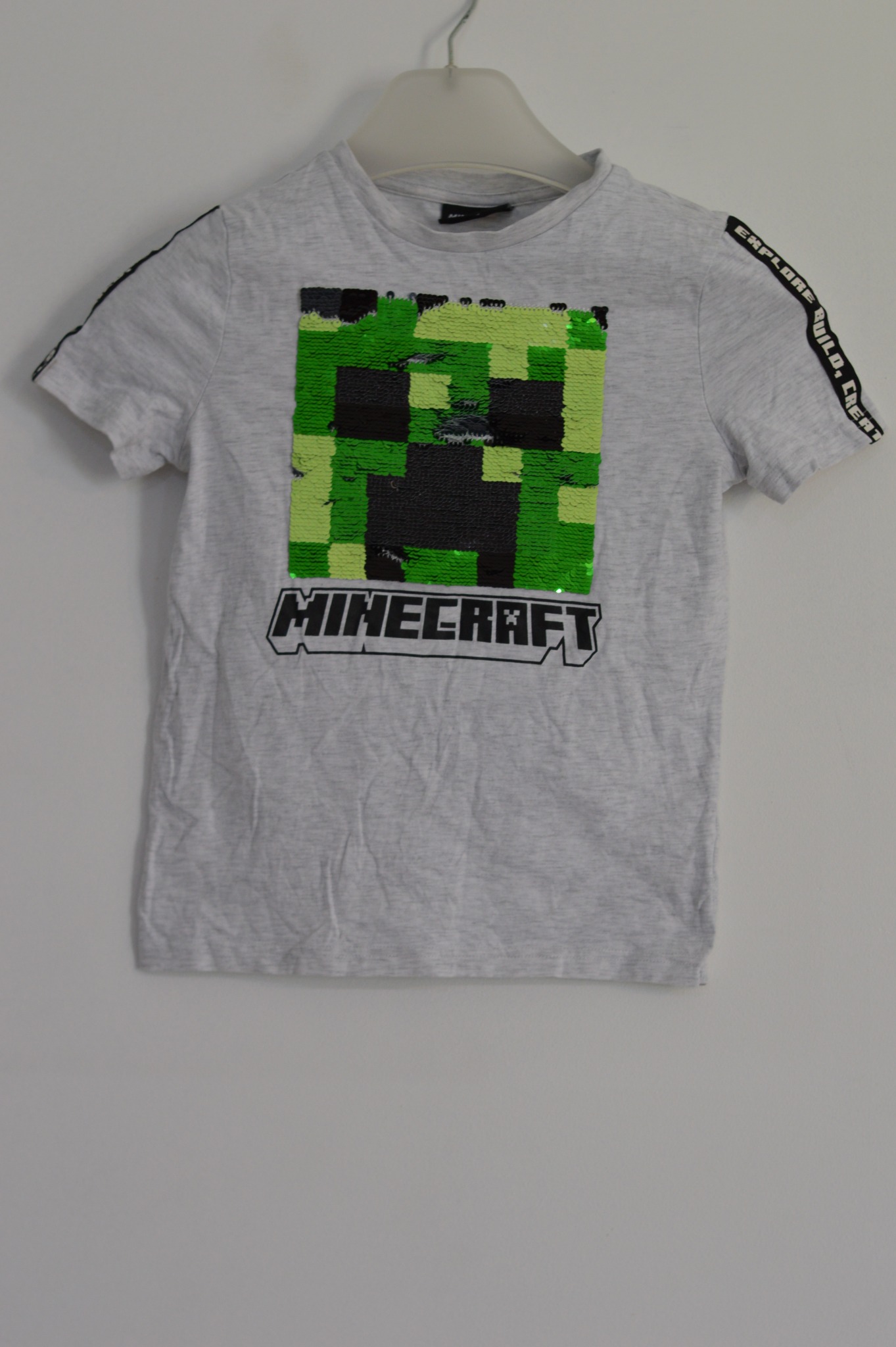 Szara koszulka z krótkim rękawem, z cekinowym motywem pikselowego ludzika z gry Minecraft i napisem 'Minecraft', wisząca na plastikowym wieszaku.