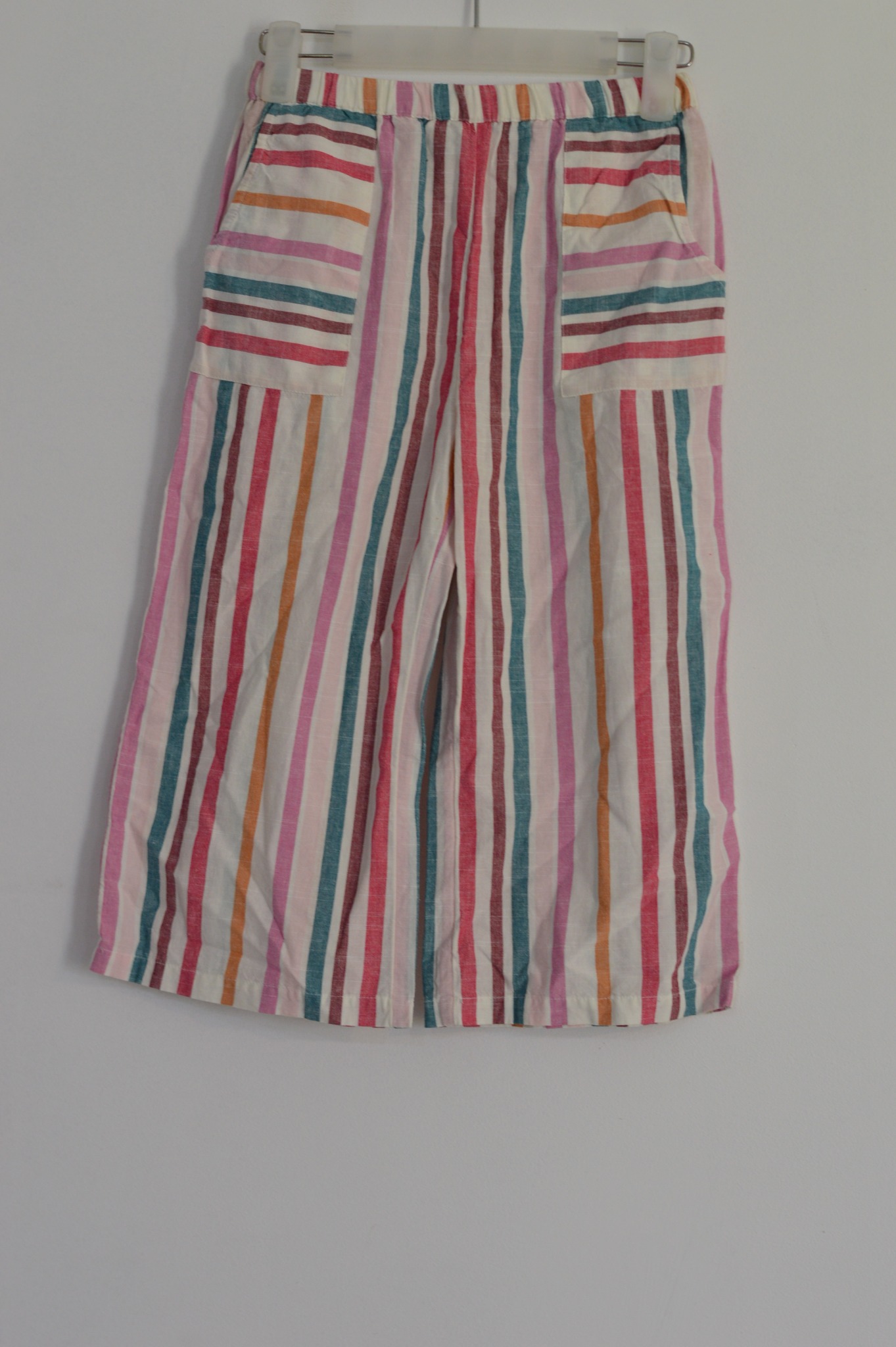 Szerokie spodnie w kolorowe pionowe paski, wiszące na wieszaku na białym tle, z widocznymi kieszeniami z poziomymi paskami.