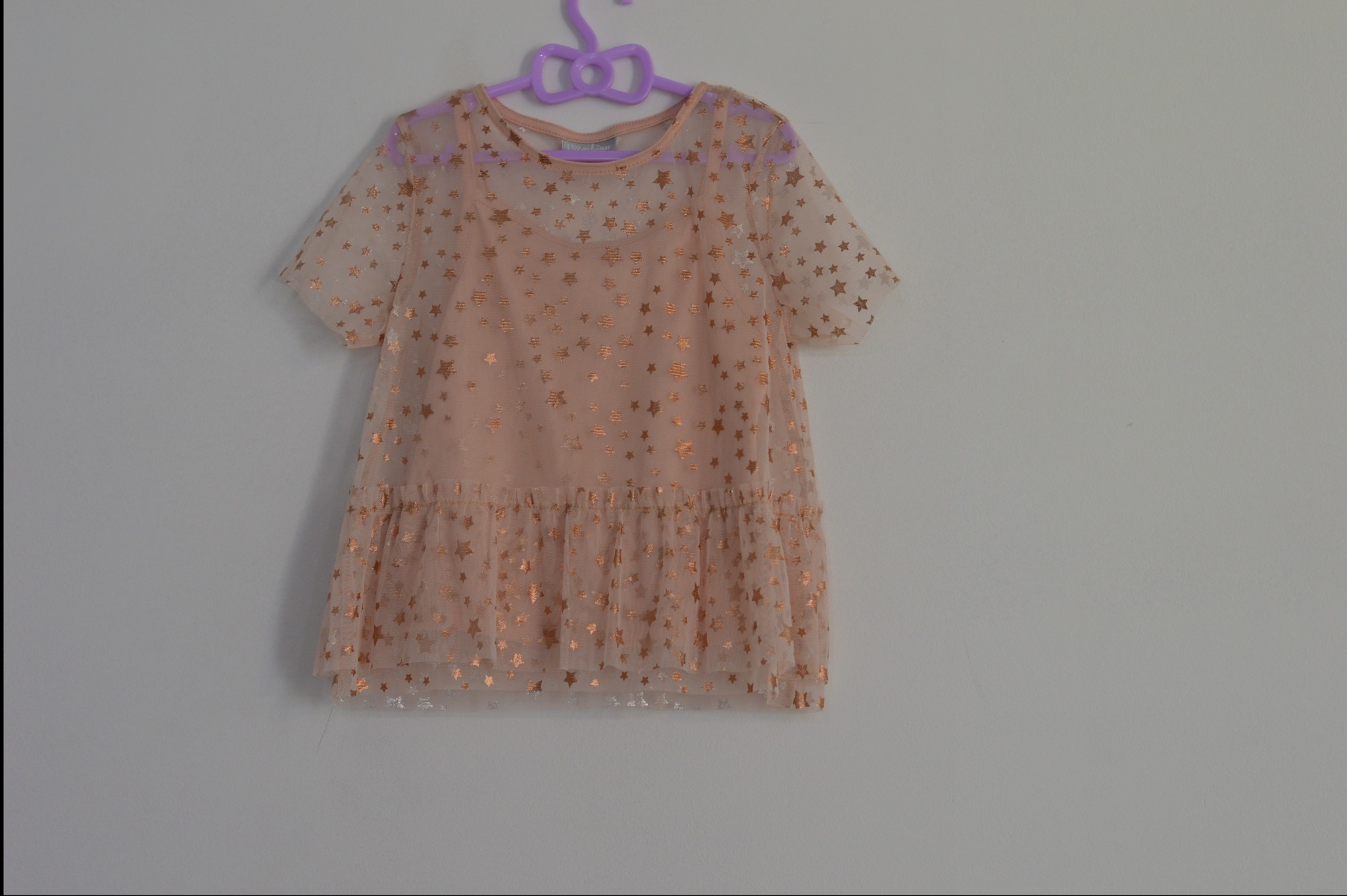 Delikatna, beżowa sukienka z tiulu w złote gwiazdki, zawieszona na fioletowym wieszaku z kokardą, na tle jasnej ściany.