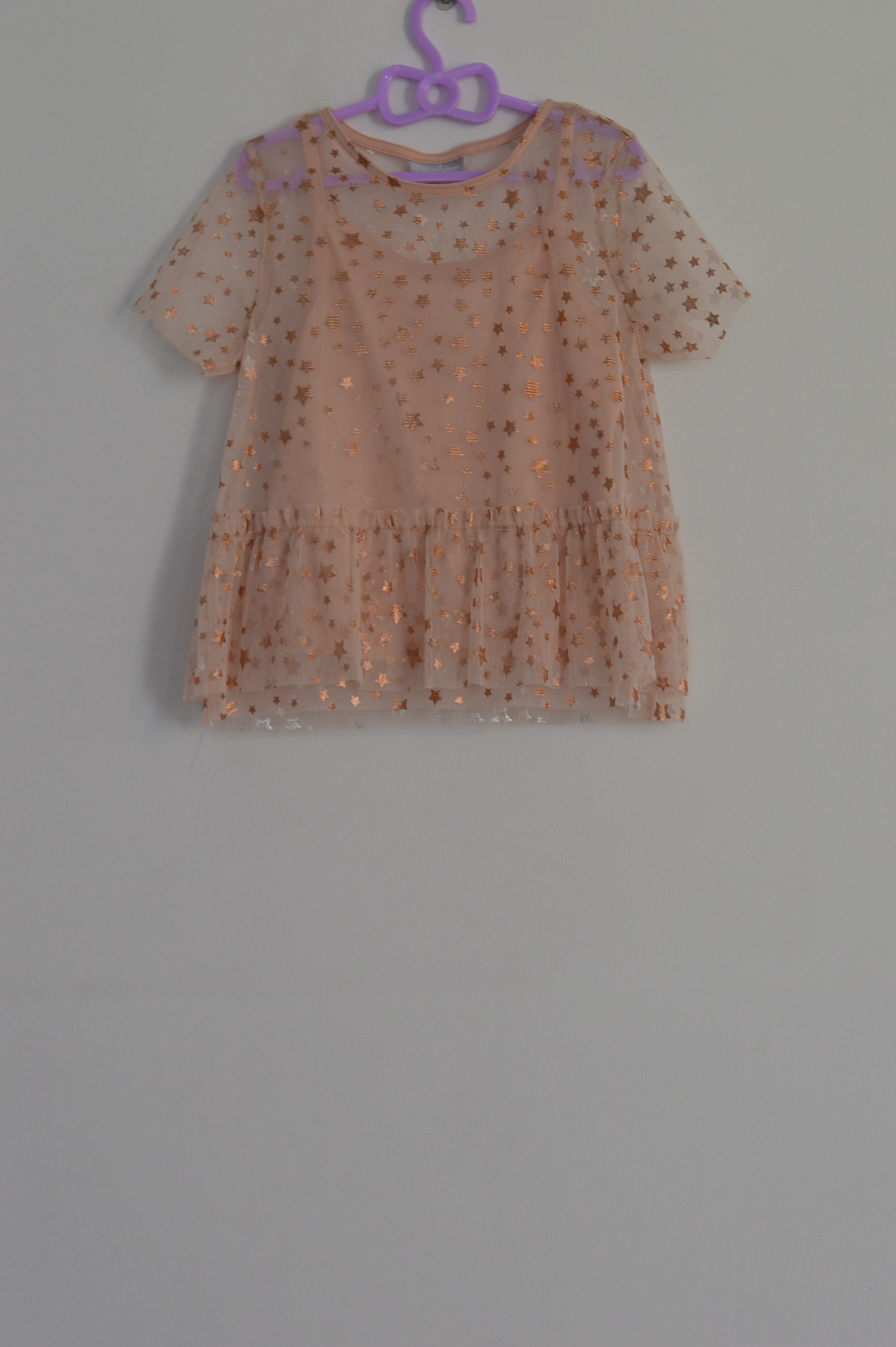 Delikatna, beżowa sukienka z tiulu, zdobiona złotymi gwiazdkami, wisząca na fioletowym wieszaku na tle białej ściany.