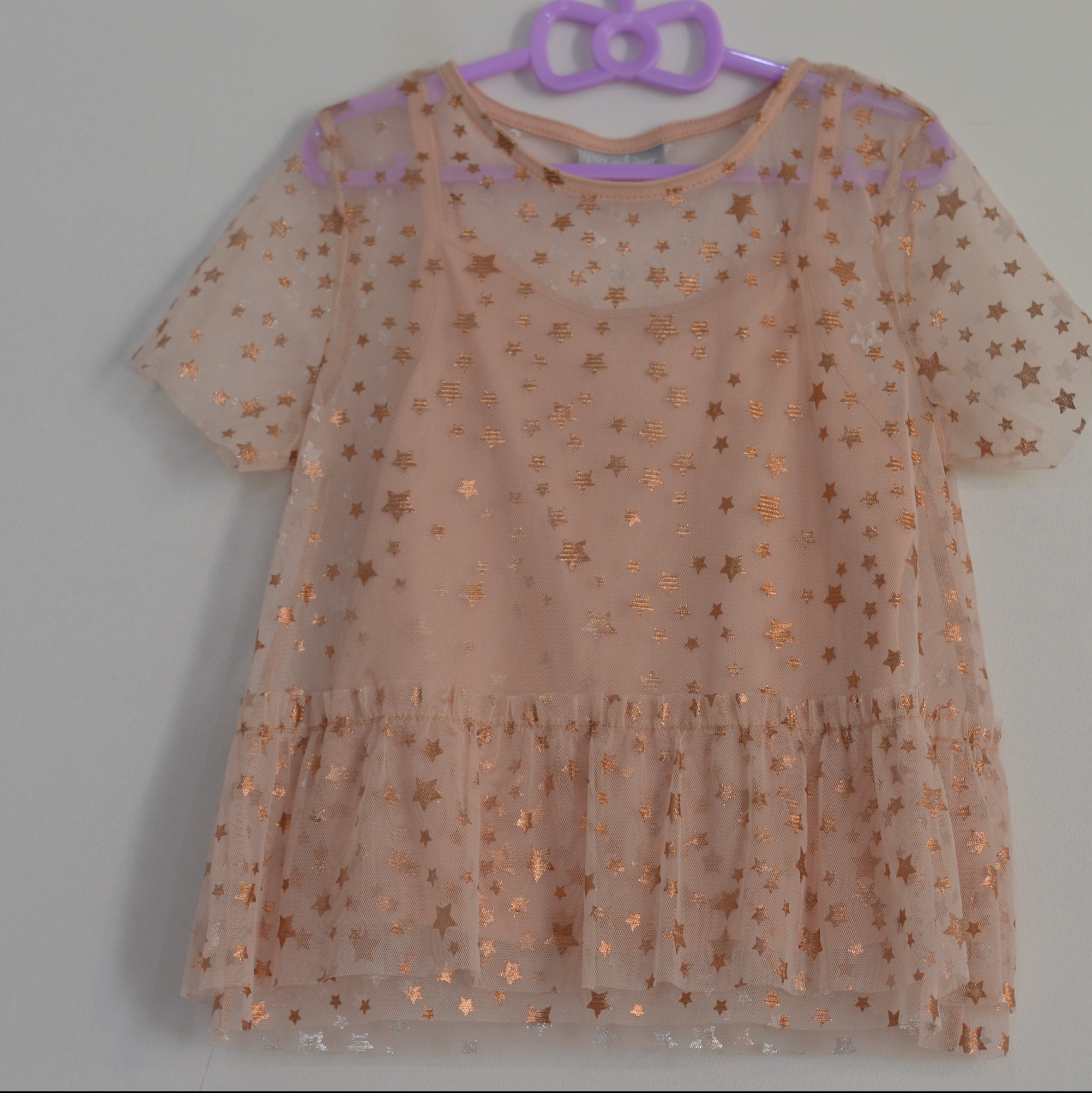 Delikatna, beżowa bluzka z tiulu w złote gwiazdki, zawieszona na fioletowym wieszaku w kształcie kokardki.