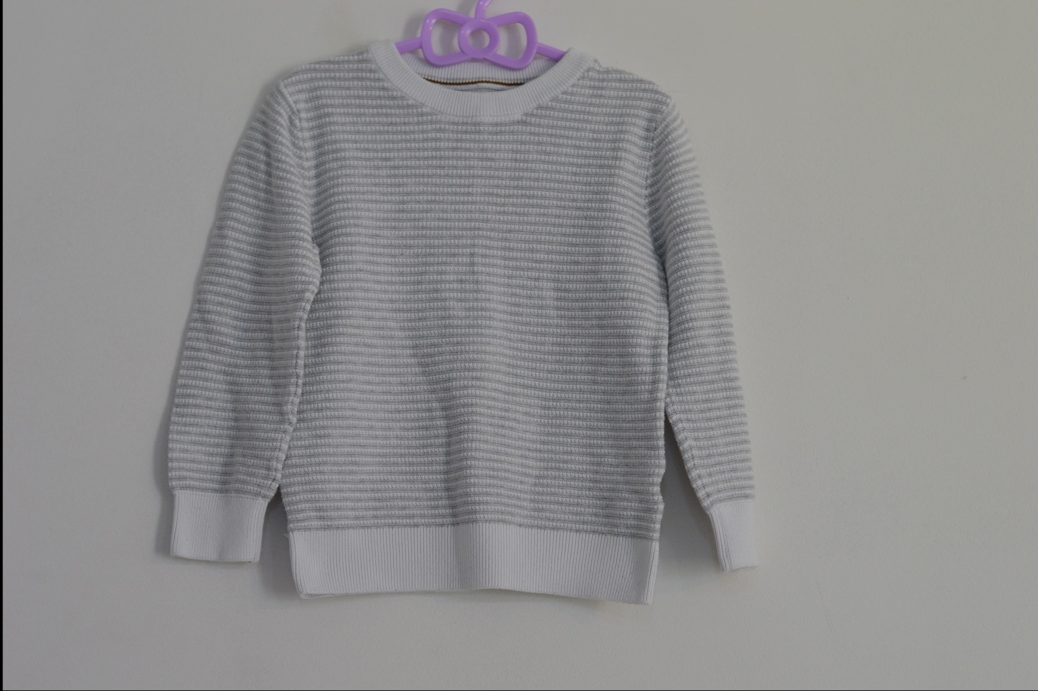 Szaro-biały sweter w poziome paski na fioletowym wieszaku, wiszący na białej ścianie.