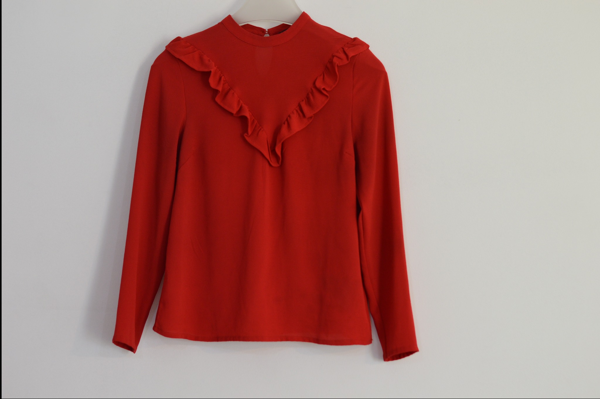 Czerwona bluzka z długim rękawem i falbanami przy dekolcie, wisząca na wieszaku na białym tle.