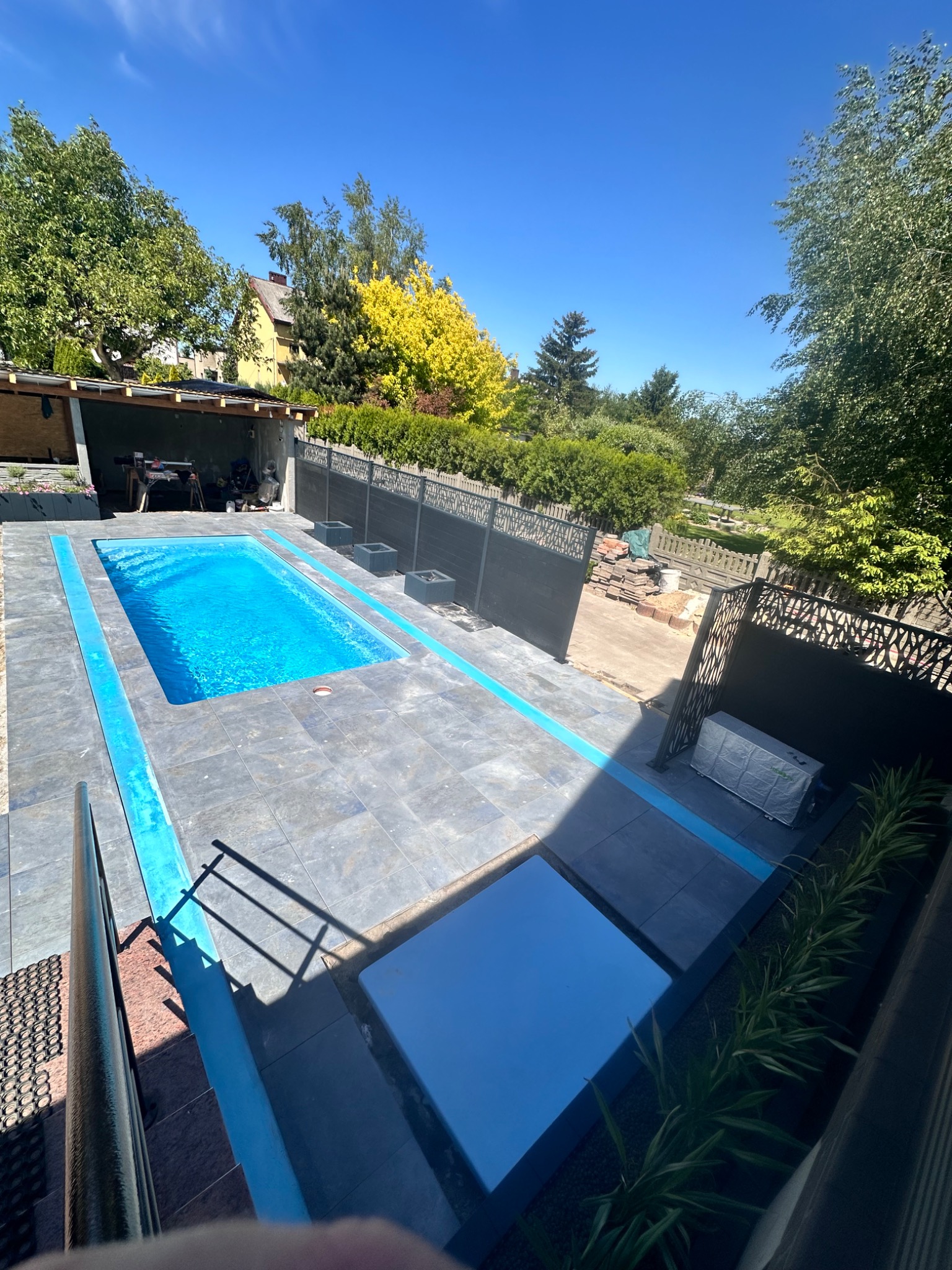 Niebieski basen otoczony szarą kostką brukową z niebieskimi akcentami, widok z góry, z elementami dekoracyjnymi i zielenią w tle.