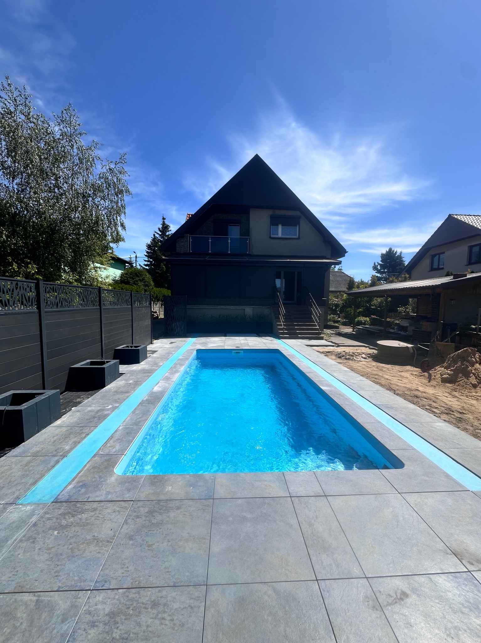 Nowoczesny basen otoczony szarą kostką, z widocznym domem i elementami ogrodu w tle, błękitna woda w basenie odbija niebo.