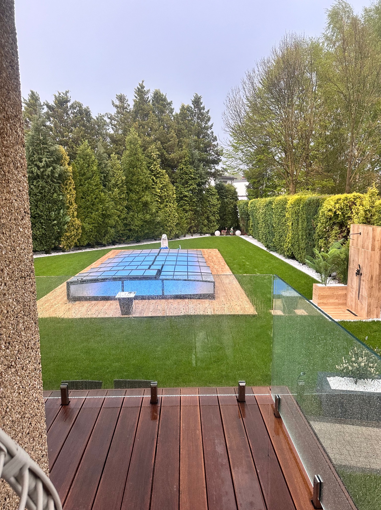 Widok na zadaszony basen z perspektywy drewnianego tarasu z szklaną balustradą, otoczony zielonym trawnikiem, strzyżonymi tujami i prysznicem ogrodowym z drewnianą obudową.