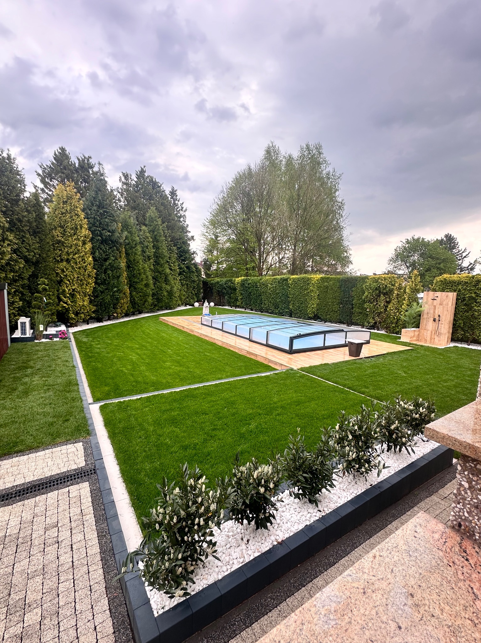 Zadbany ogród z równo przystrzyżonym trawnikiem, basenem z zadaszeniem, obrzeżami z kostki brukowej i białymi kamieniami dekoracyjnymi pod krzewami.