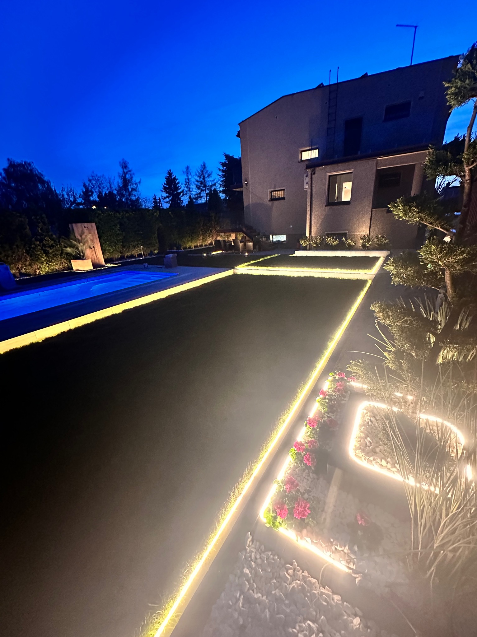 Oświetlenie dekoracyjne ogrodu z podświetlonym basenem i rabatami kwiatowymi, widoczne wieczorem na tle domu.
