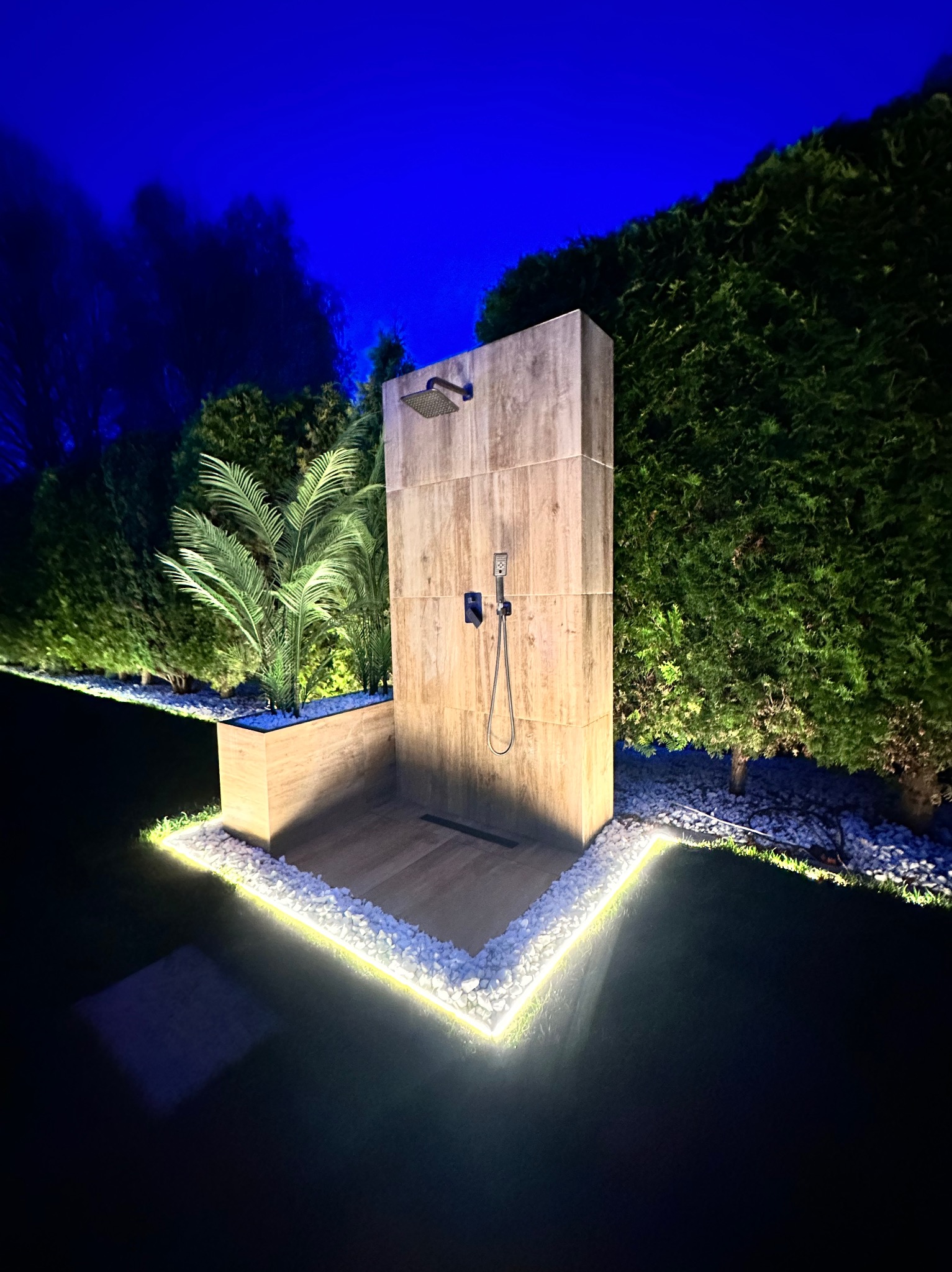 Oświetlony zewnętrzny prysznic z kamienną obudową i deszczownicą, otoczony ozdobnymi kamieniami i zielenią w nocnym oświetleniu.
