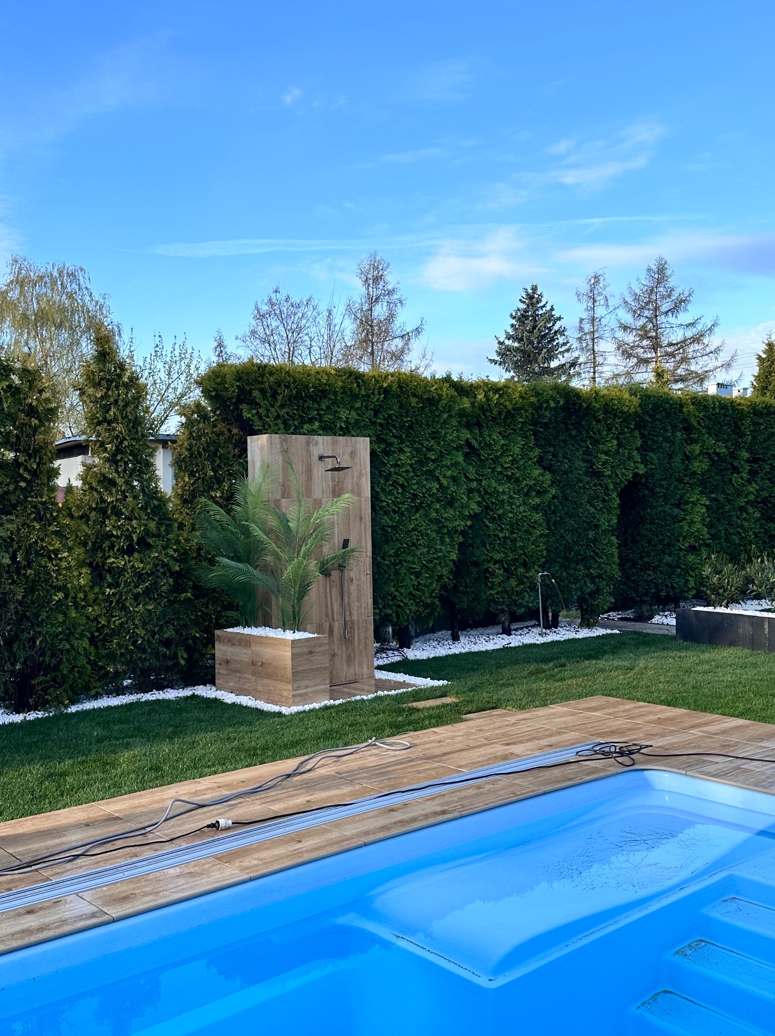 Nowoczesny prysznic ogrodowy z drewnopodobnych płyt, obok basenu, otoczony zielenią i białym żwirem, na tle żywopłotu.