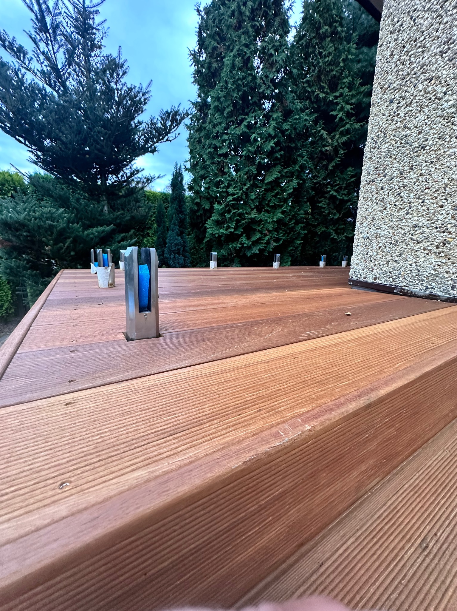 Drewniany taras z zamontowanymi stalowymi uchwytami z niebieskim wypełnieniem, przygotowany pod montaż szklanej balustrady, widok z niskiej perspektywy.