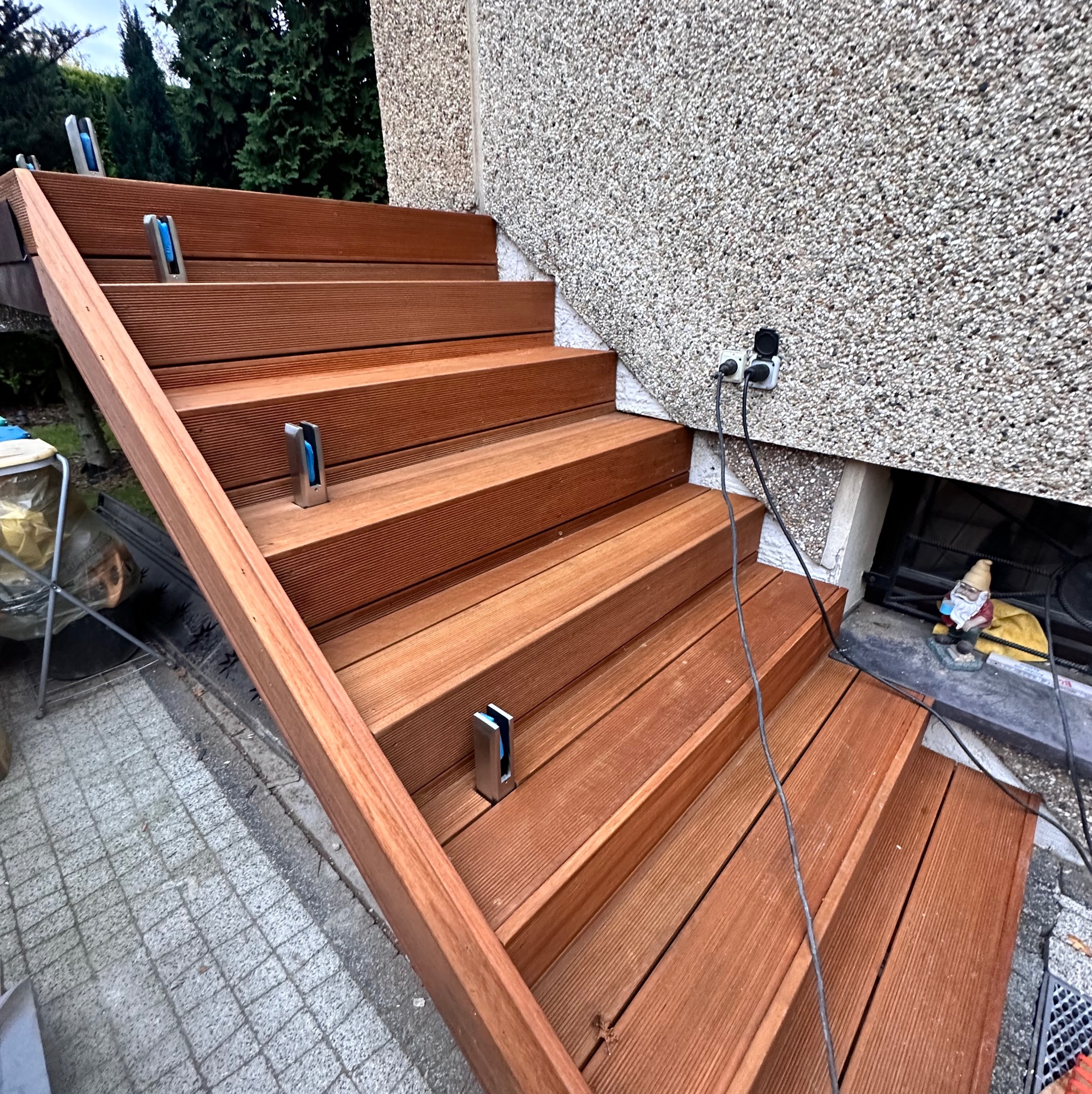 Drewniane schody zewnętrzne z metalowymi uchwytami na balustradę szklaną, zamontowane przy elewacji budynku z gniazdkami elektrycznymi i dekoracyjnym krasnalem w piwnicy.