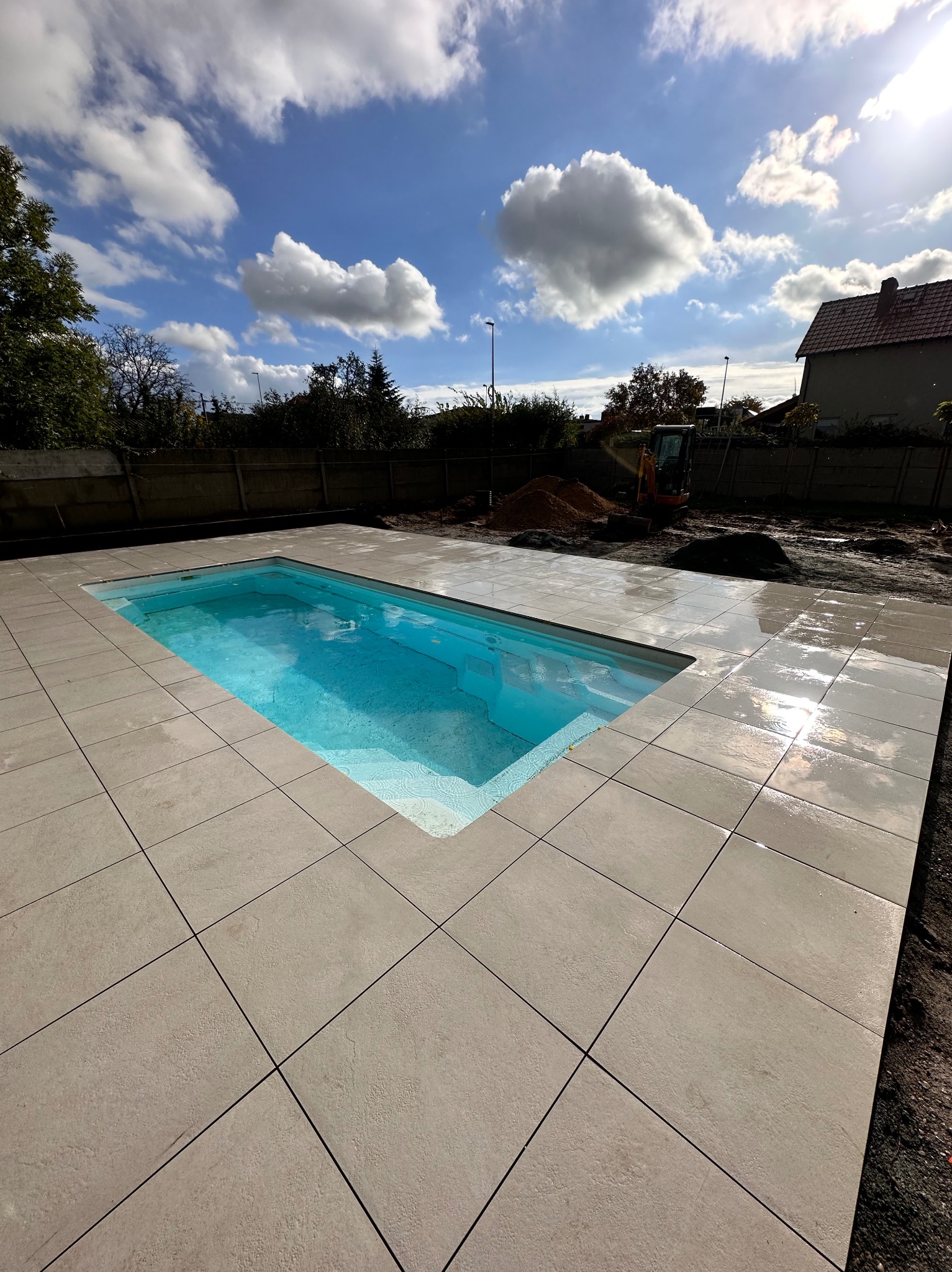 Nowoczesny basen z turkusową wodą otoczony jasnymi, kwadratowymi płytami chodnikowymi; w tle widoczny mały koparka i błękitne niebo z chmurami.