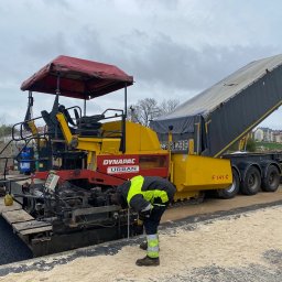 Piotr Urban - Układanie asfaltu za pomocą rozściełacza Dynapac Urban i ciężarówki Wielton. Pracownik w odblaskowej kamizelce kontroluje proces. Widoczny świeży asfalt i przygotowane podłoże.