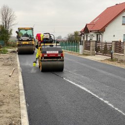Piotr Urban - Drogowcy w żółtych kamizelkach układają asfalt na ulicy, używając walców drogowych marki Urban Dynapac, w tle widoczny dom z czerwonym dachem i ogrodzeniem.