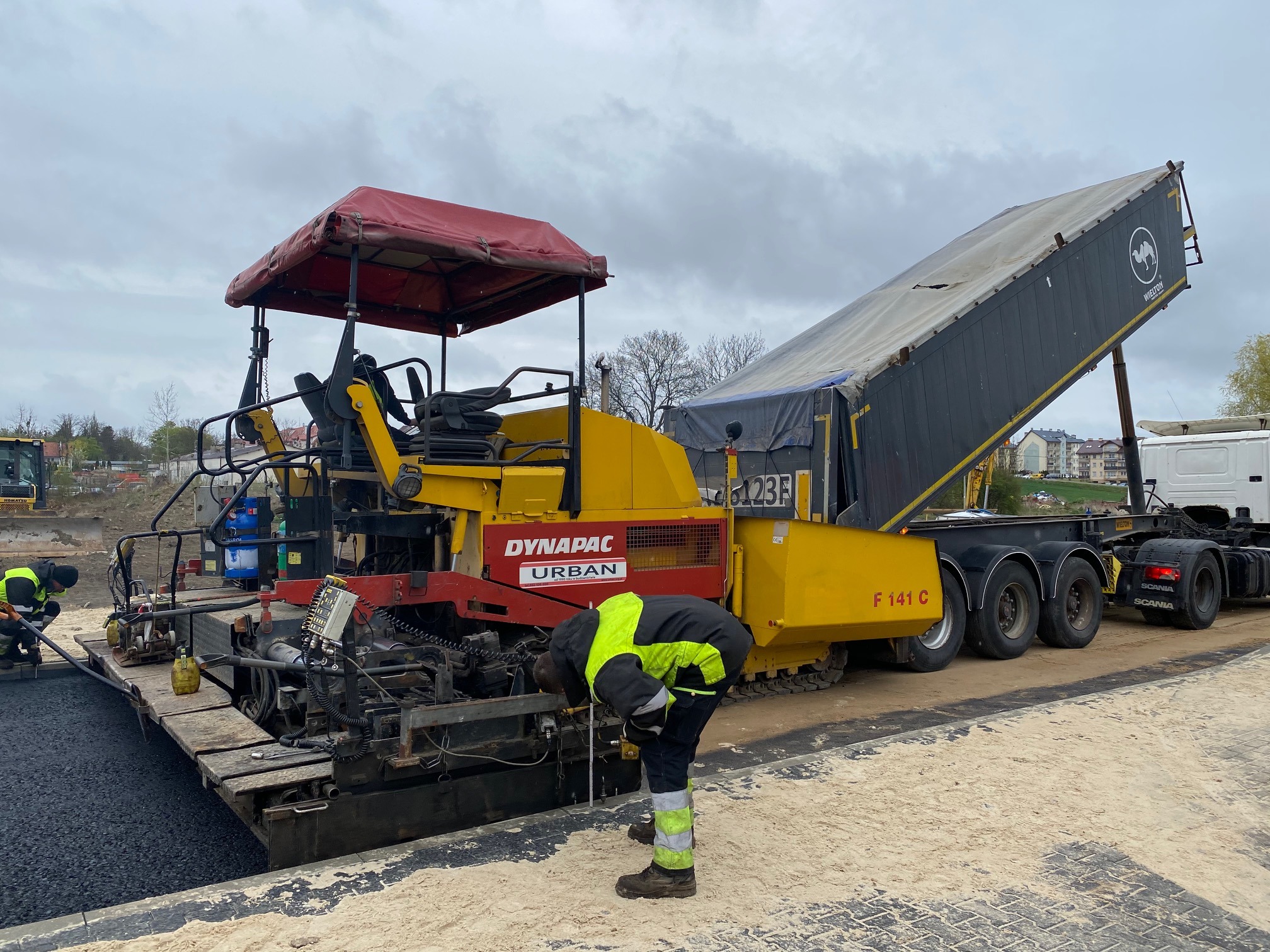 Układanie asfaltu za pomocą rozściełacza Dynapac Urban i ciężarówki Wielton. Pracownik w odblaskowej kamizelce kontroluje proces. Widoczny świeży asfalt i przygotowane podłoże.