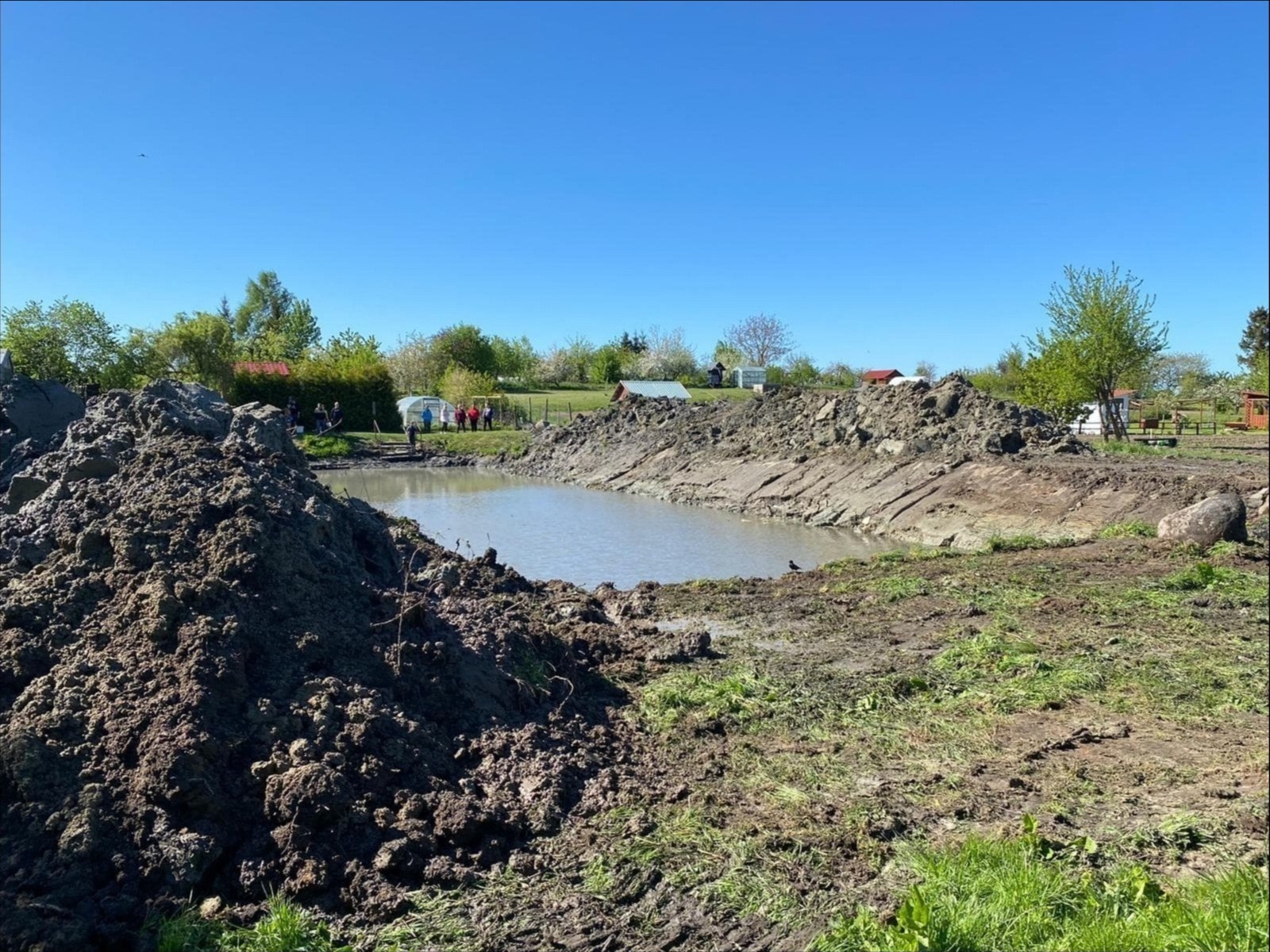 Świeżo wykopany staw wypełniony wodą, otoczony stertami ziemi i zielenią, z grupą osób obserwujących na dalszym planie w słoneczny dzień.