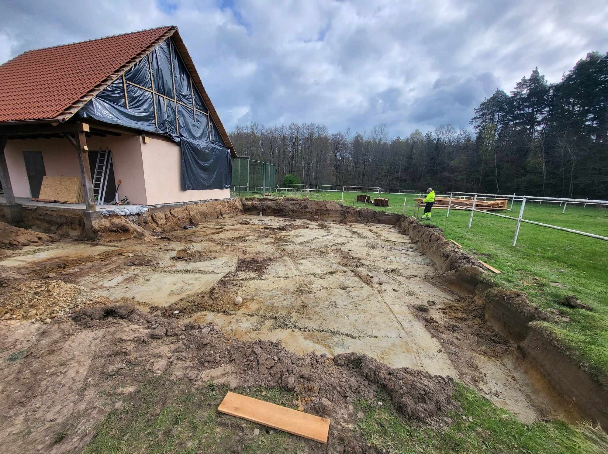Szeroki wykop pod fundamenty domu z częściowo zakrytą plandeką elewacją, widoczny pracownik w odblaskowej kamizelce i ułożone drewniane elementy na trawiastym terenie.