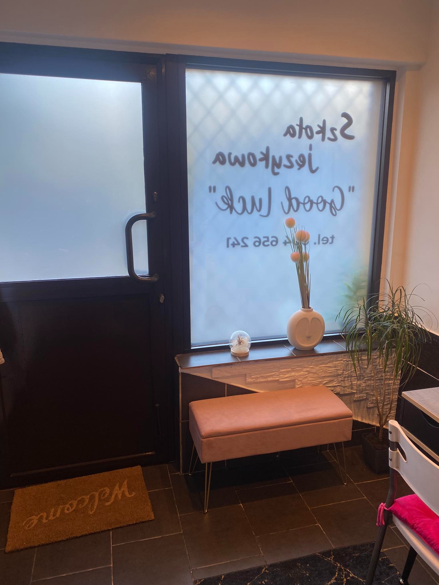 Wejście do szkoły językowej z napisem 'Good Luck' na matowej szybie, obok stoi ławka z weluru na złotych nóżkach i doniczka z zieloną rośliną.