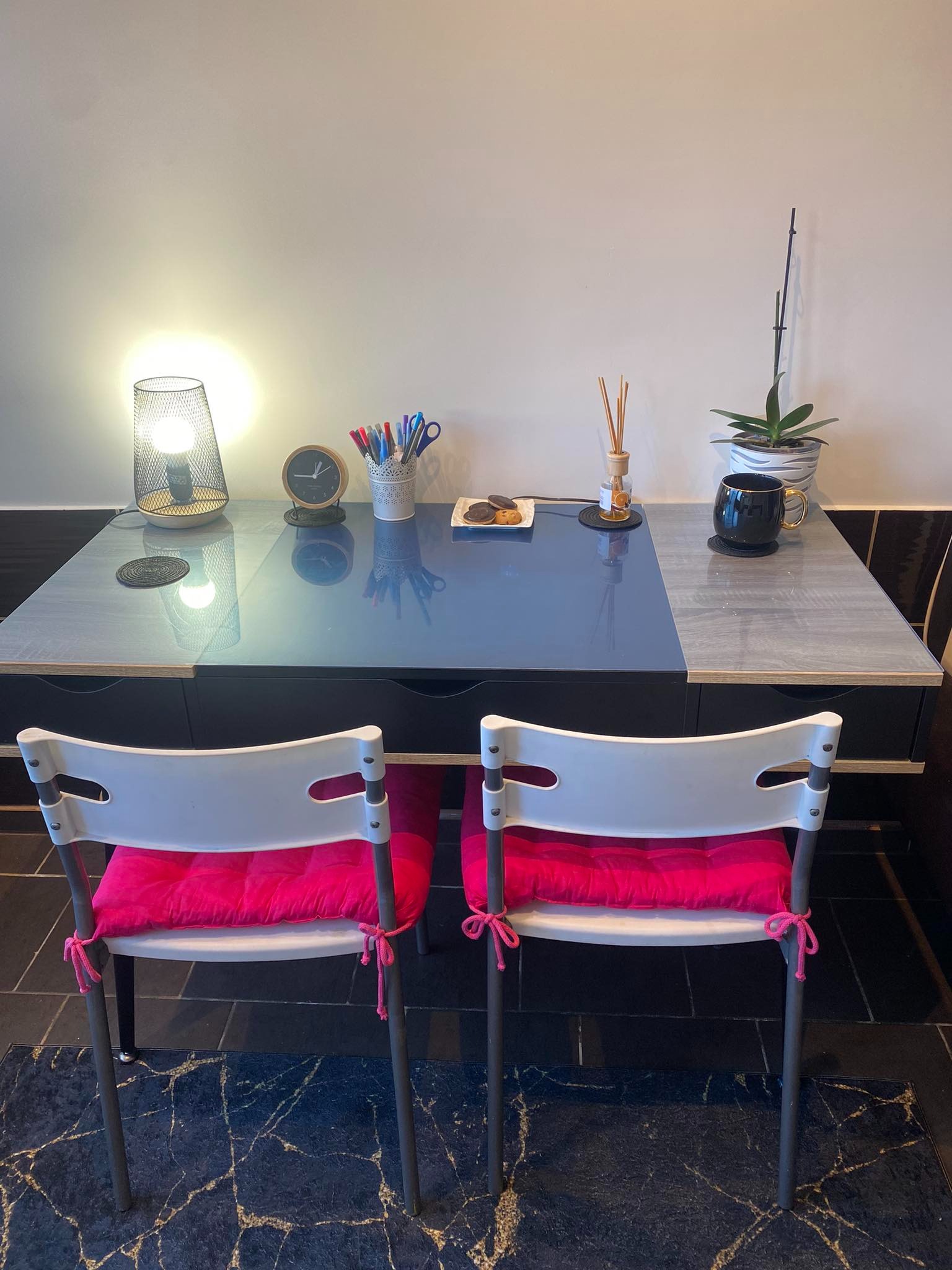 Stół z dwoma krzesłami z różowymi poduszkami, lampka nocna, zegar, przybory do pisania, ciastka, dyfuzor zapachowy i roślina doniczkowa tworzą przytulne miejsce do nauki.