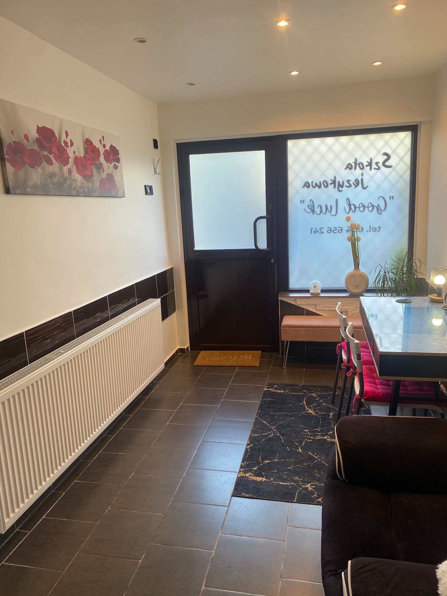 Wnętrze jasnego pokoju z czarną podłogą, białym grzejnikiem i oknem z napisem 'Good luck' po angielsku, dekoracje kwiatowe i obraz z motywem czerwonych maków na ścianie.