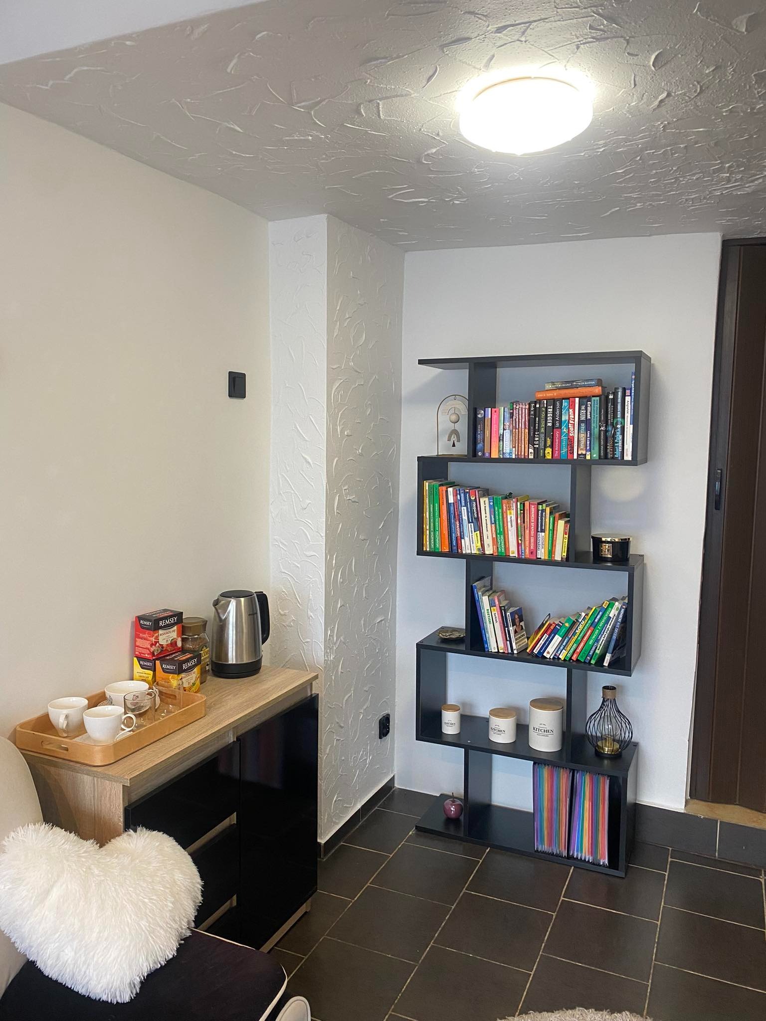 Kącik z regałem pełnym książek, czajnikiem i herbatą, prawdopodobnie w sali lekcyjnej lub gabinecie nauczyciela.