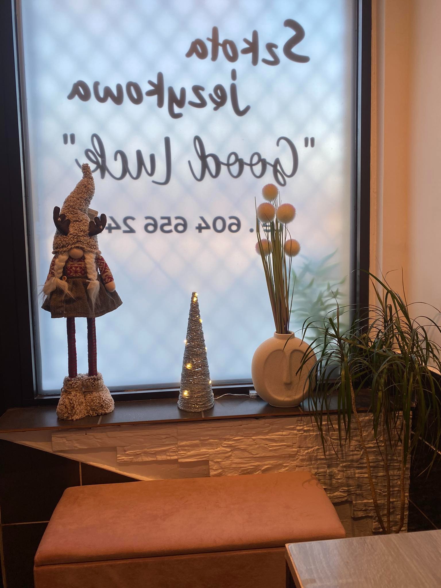 Dekoracja okienna z figurką gnoma w stroju świątecznym, małą choinką z lampkami, wazonem z kulistymi kwiatami i rośliną doniczkową na tle okna z napisem 'Good Luck' i odwróconymi słowami.