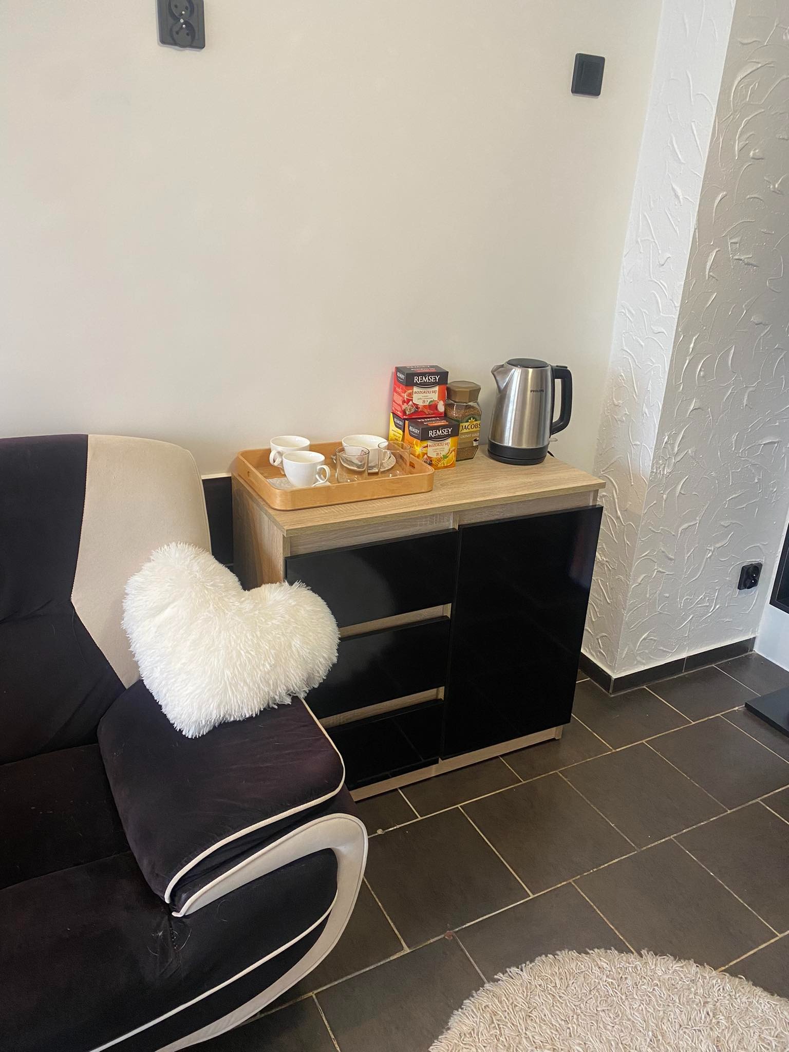 Stolik z czarnymi drzwiczkami, na nim czajnik elektryczny, pudełka herbaty i taca z filiżankami, obok czarna kanapa z białą poduszką w kształcie serca.