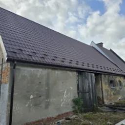 Dwuspadowy dach pokryty grafitową blachodachówką łuskową na budynku w trakcie renowacji, z widocznym systemem rynnowym i obróbkami blacharskimi w tym samym kolorze, rusztowaniem przy ścianie...