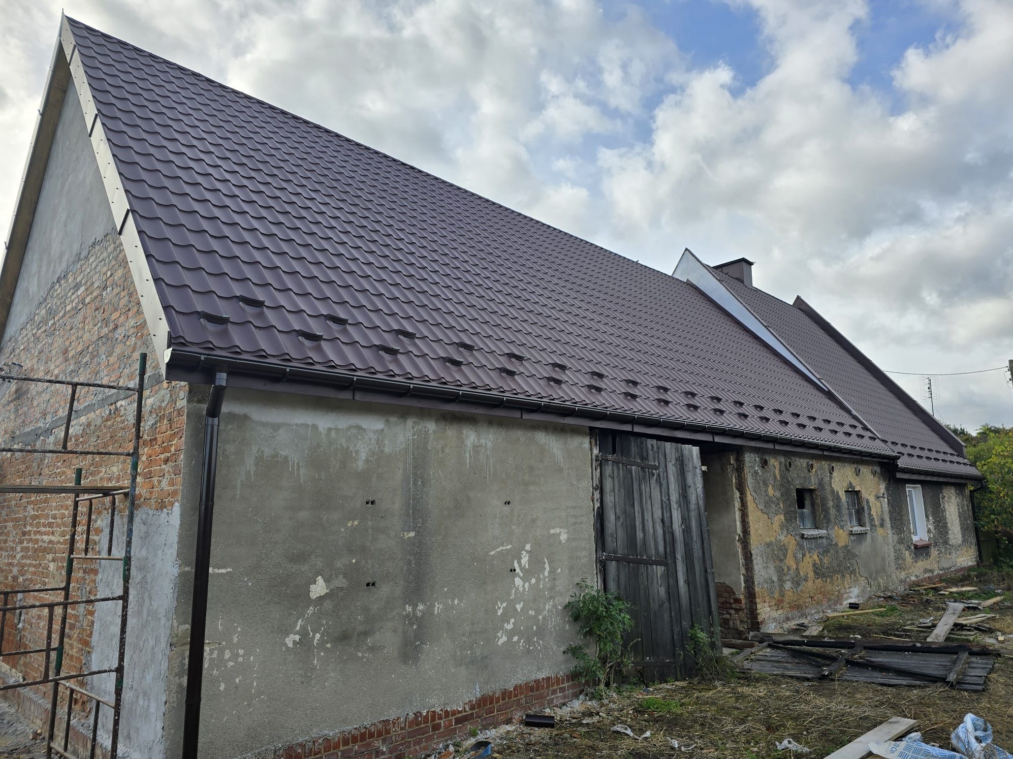 Dwuspadowy dach pokryty grafitową blachodachówką łuskową na budynku w trakcie renowacji, z widocznym systemem rynnowym i obróbkami blacharskimi w tym samym kolorze, rusztowaniem przy ścianie...