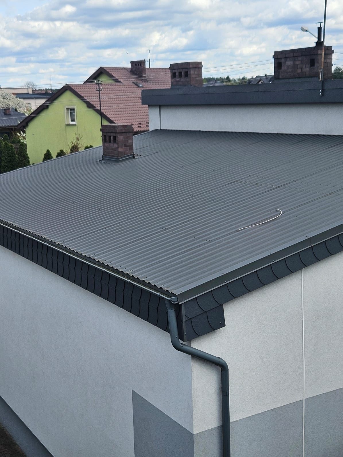 Grafitowa blacha trapezowa na dachu budynku z widocznymi obróbkami blacharskimi, pasem nadrynnowym i narożnikami w tym samym kolorze, z rynną spustową. W tle widoczne inne budynki i pochmurne niebo.