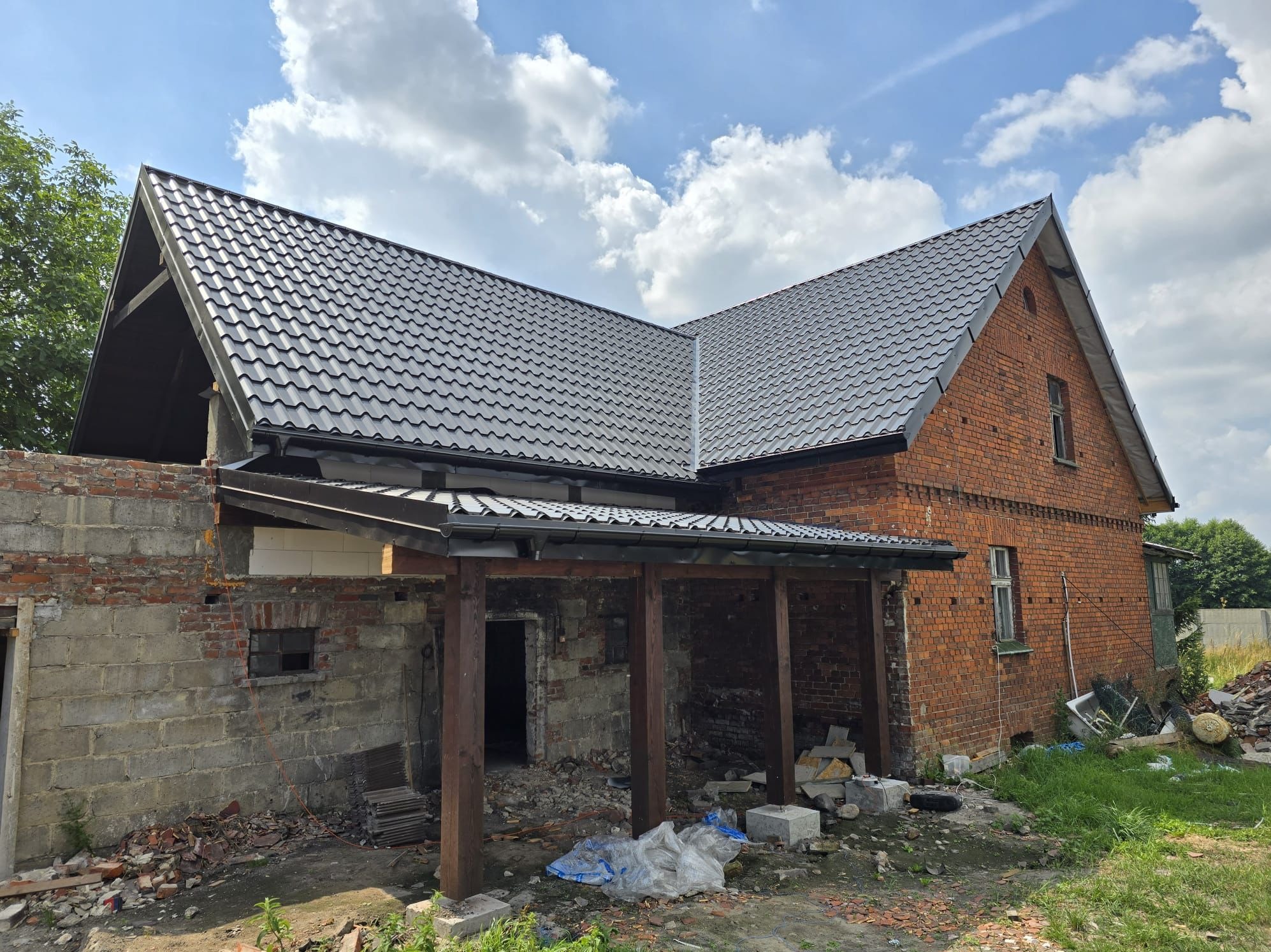 Grafitowy dach wielospadowy z blachodachówki łuskowej na ceglanym budynku z widocznymi obróbkami blacharskimi, częściowo wykończony, z dobudowaną drewnianą konstrukcją i elementami gruzu na ziemi.