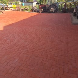 Tomasz Iwański Usługi Ogólnobudowlane - Czerwona kostka brukowa ułożona na posesji, z traktorem i żółtym pojazdem w tle, widoczne budynki i drzewa, słoneczny dzień.