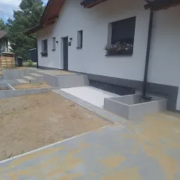 Nowoczesny dom z szarą kostką brukową, schody z betonowych bloczków i rabata z białym żwirem. Prace brukarskie w toku, widoczny piasek na kostce. Słoneczny dzień.